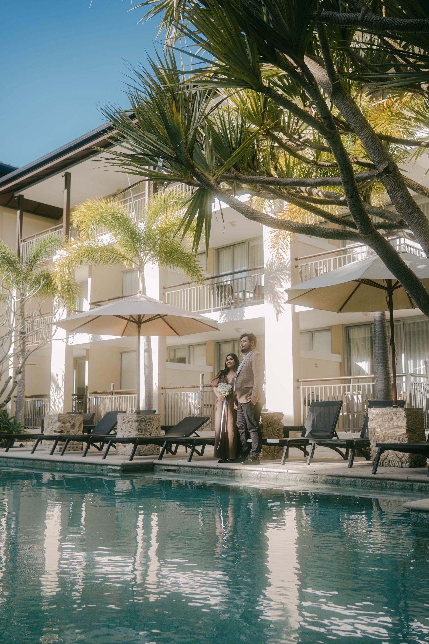 Luxurious resort swimming pool surrounded by palm trees and lounge chairs with white columns reflecting in blue water.