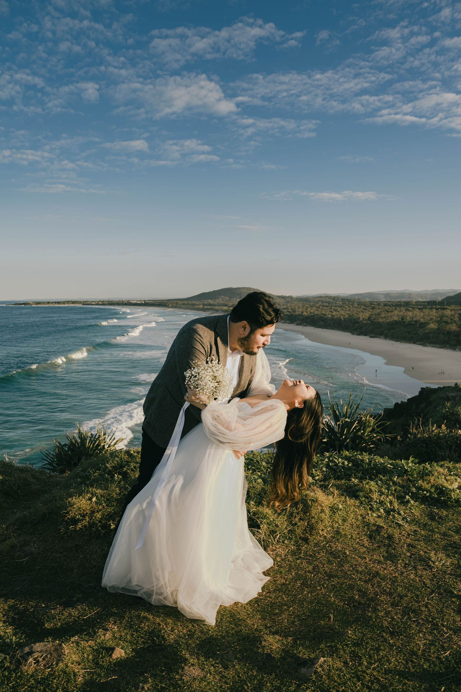 A couple in wedding attire shares intimate moments along a scenic coastal clifftop overlooking rolling waves and beaches.