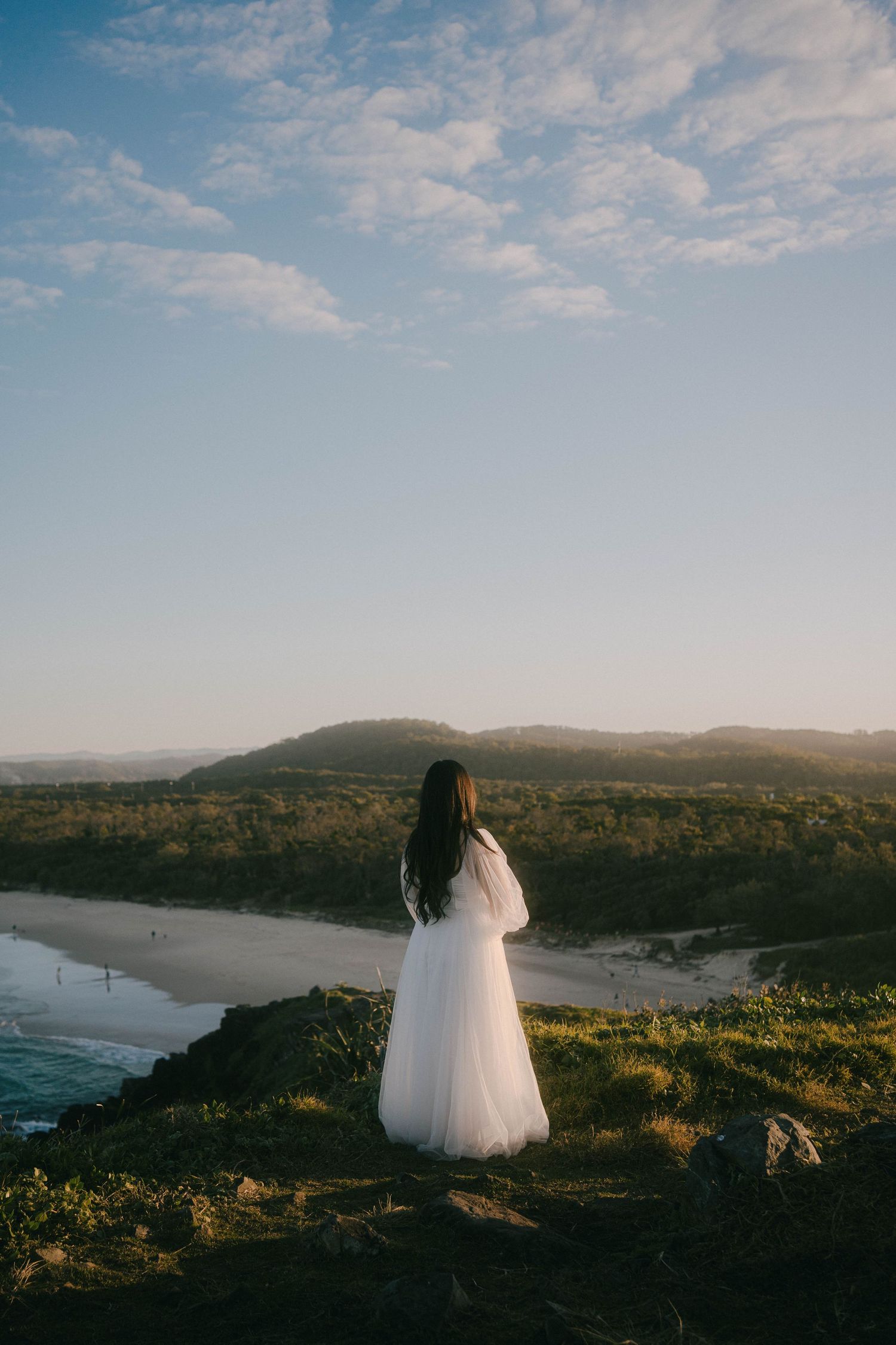 A figure in a flowing white dress stands on a cliff overlooking a scenic coastal beach at sunset.