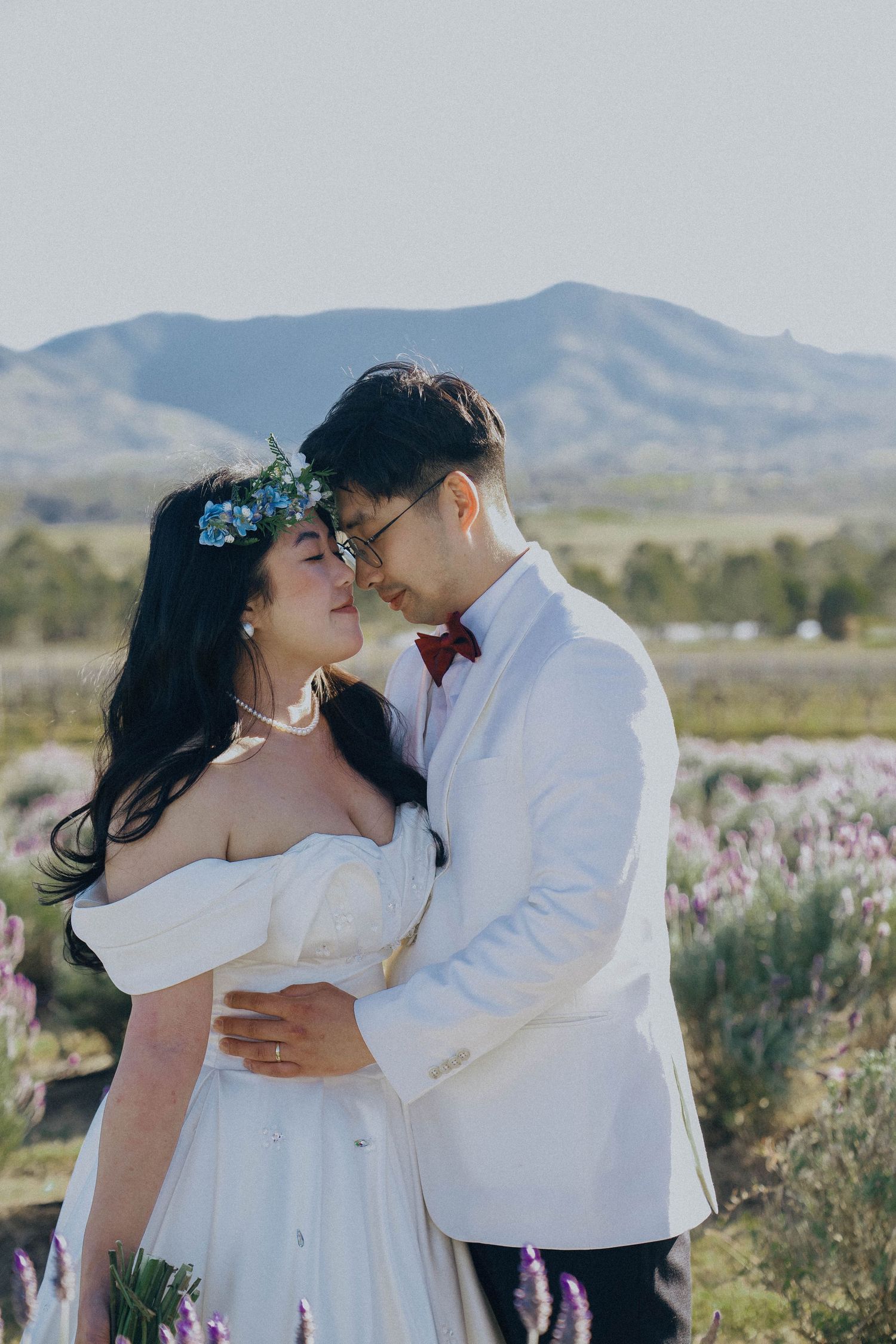 A romantic couple embraces in a field of wildflowers with mountains in the background on their wedding day.