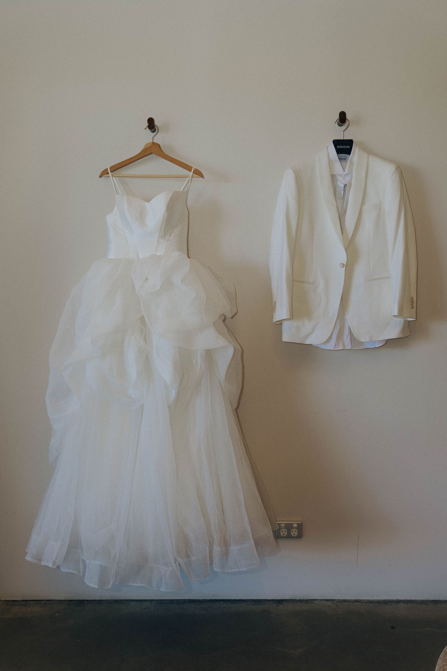 A collection of white formal garments including a wedding dress and jacket displayed on hangers against a neutral wall.