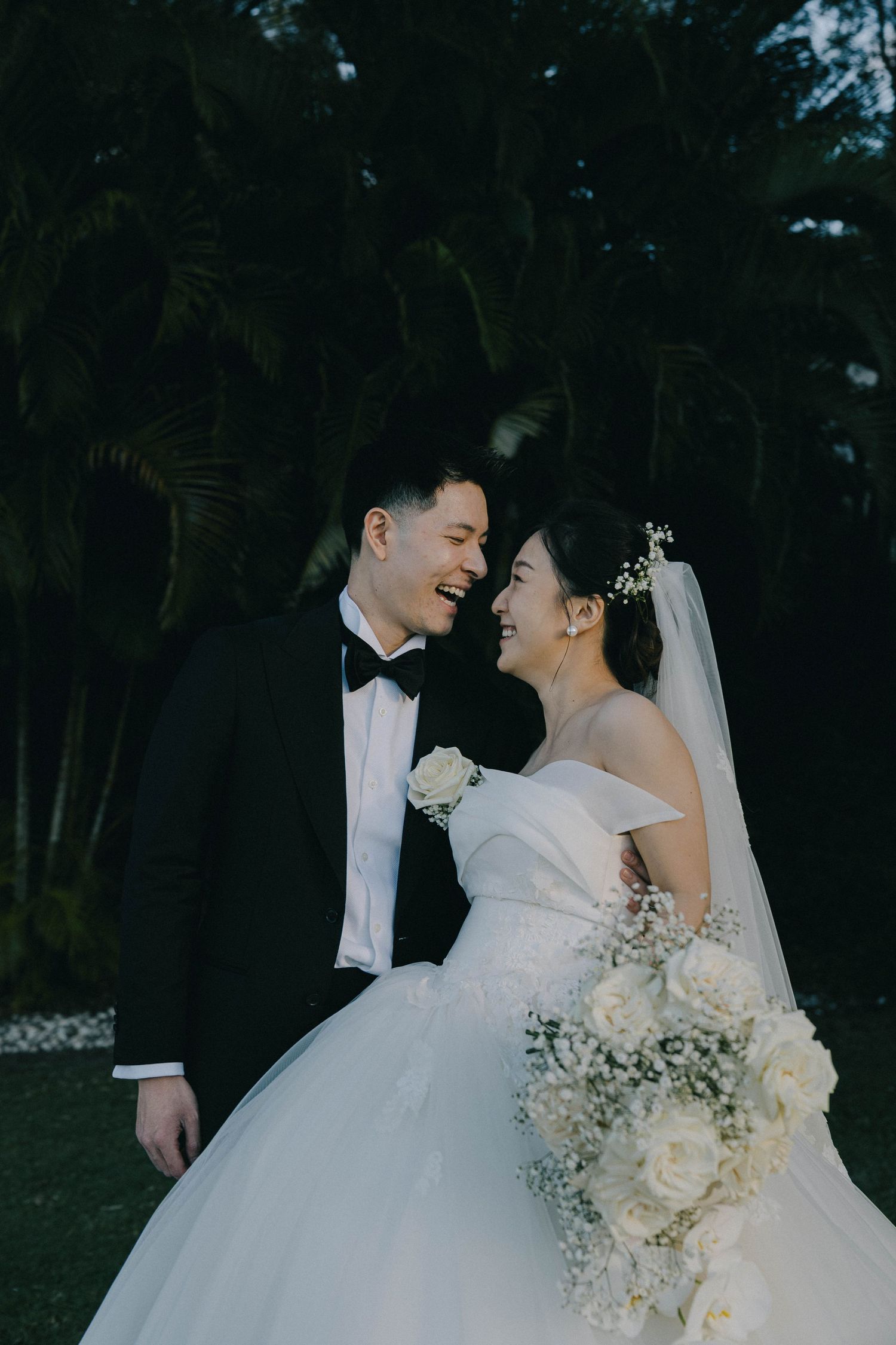 Romantic wedding couple embraces in a garden setting at dusk with delicate white floral bouquet and flowing veil.
