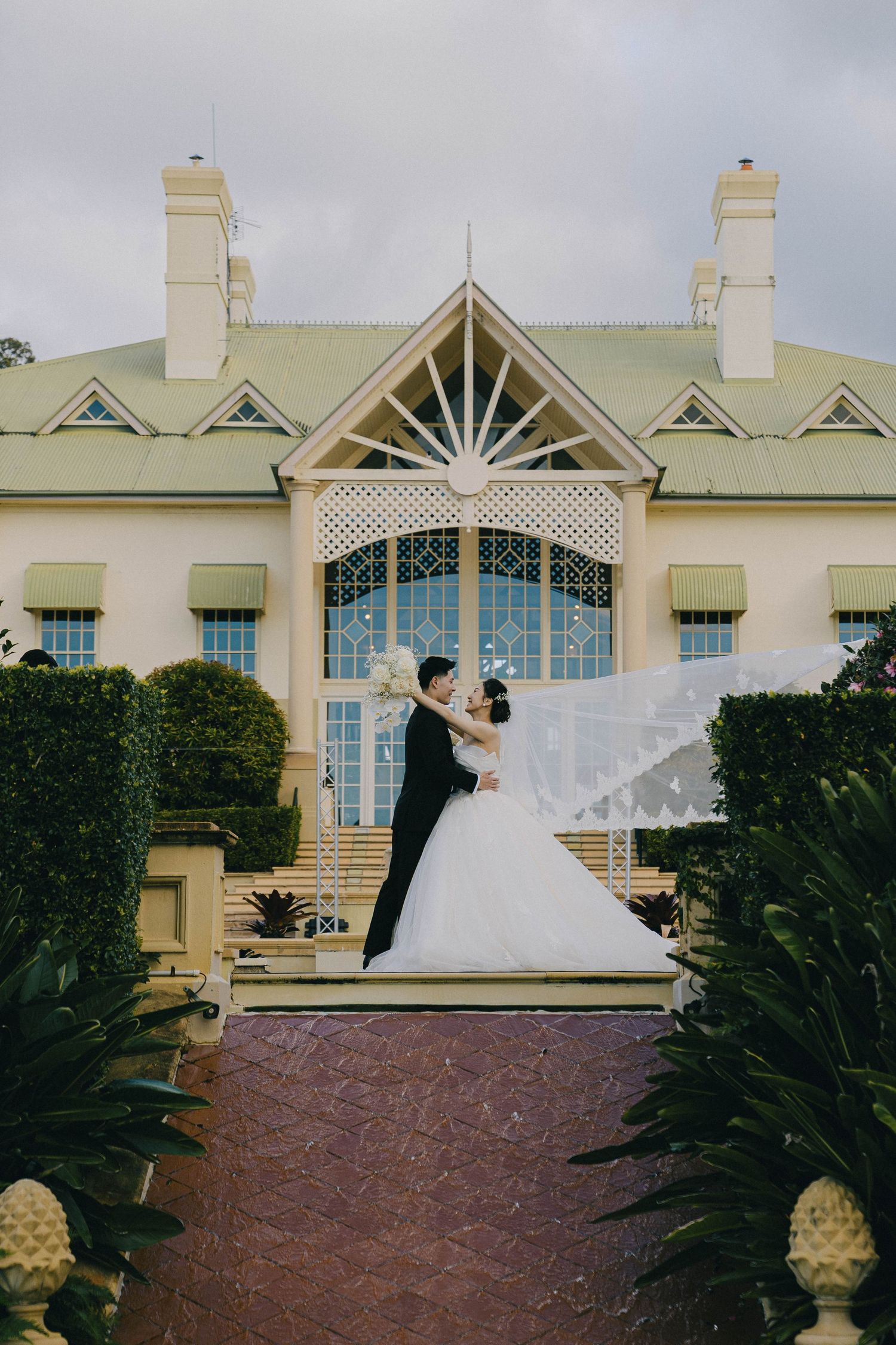 A romantic wedding couple poses against a cream colored building with ornate architectural details and tropical landscaping.