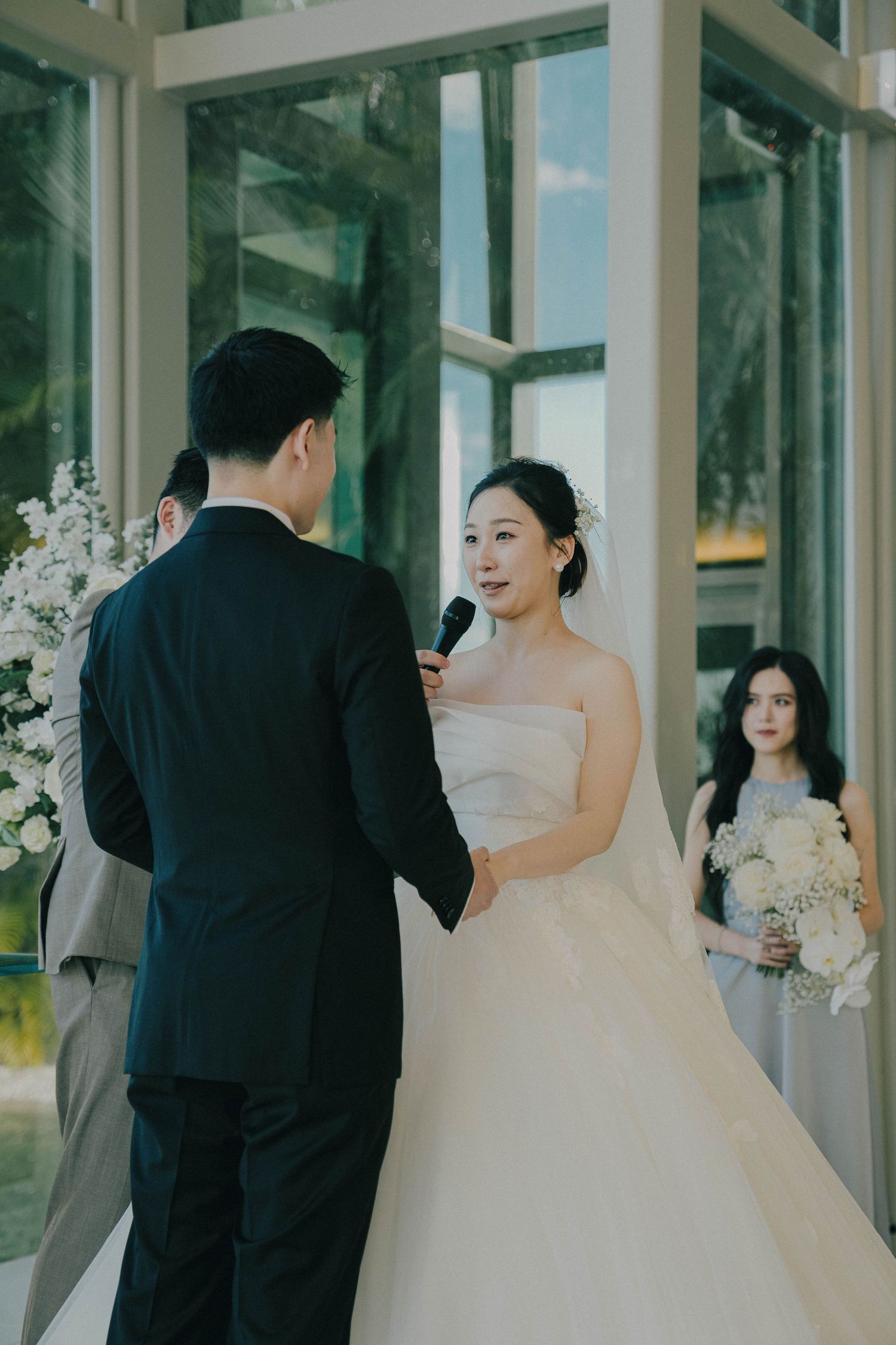 A wedding ceremony takes place in a modern glass-walled venue as the couple shares their vows with guests looking on.
