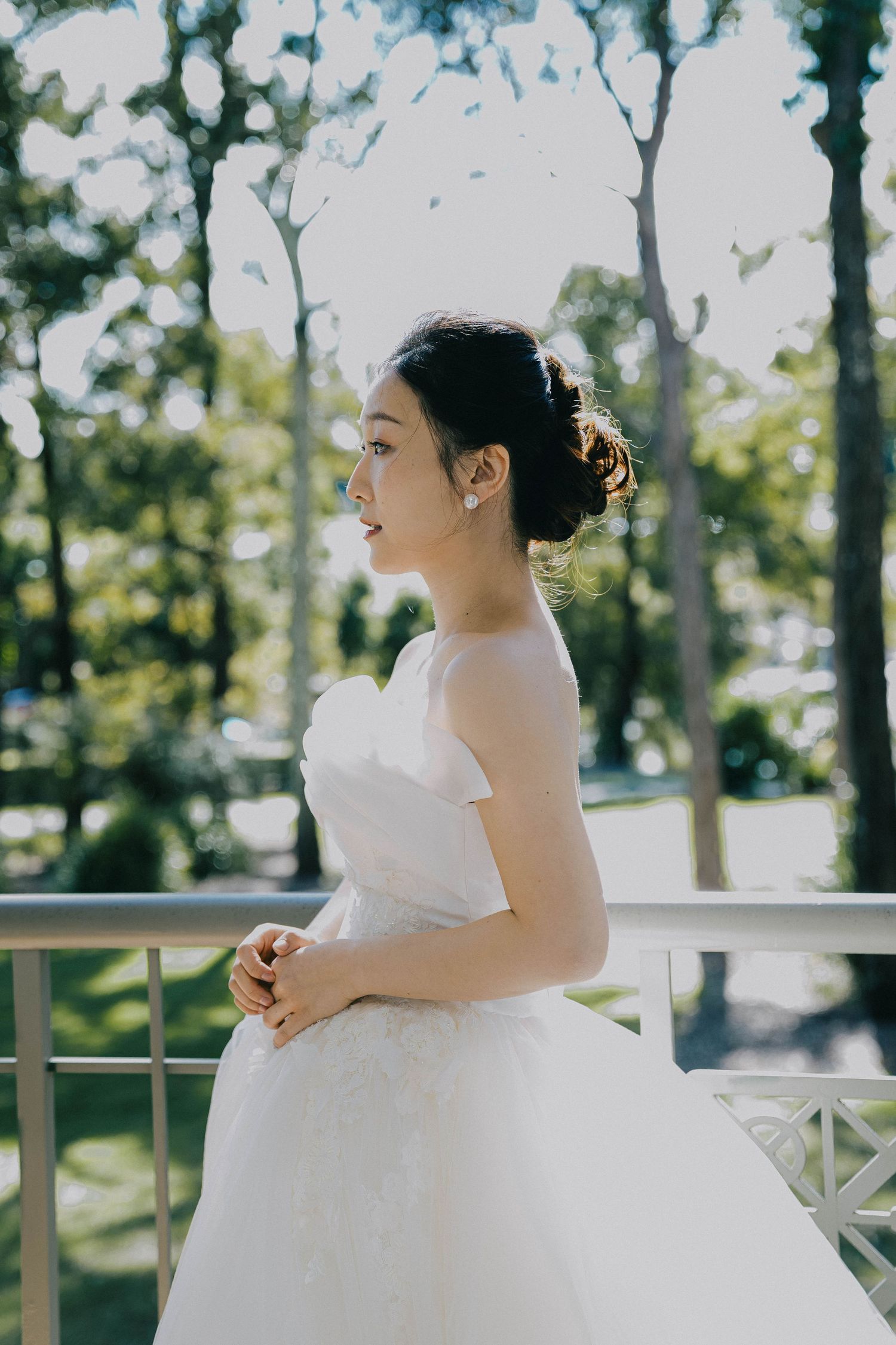 Bride in elegant white wedding gown poses gracefully on outdoor balcony surrounded by sunlit trees.