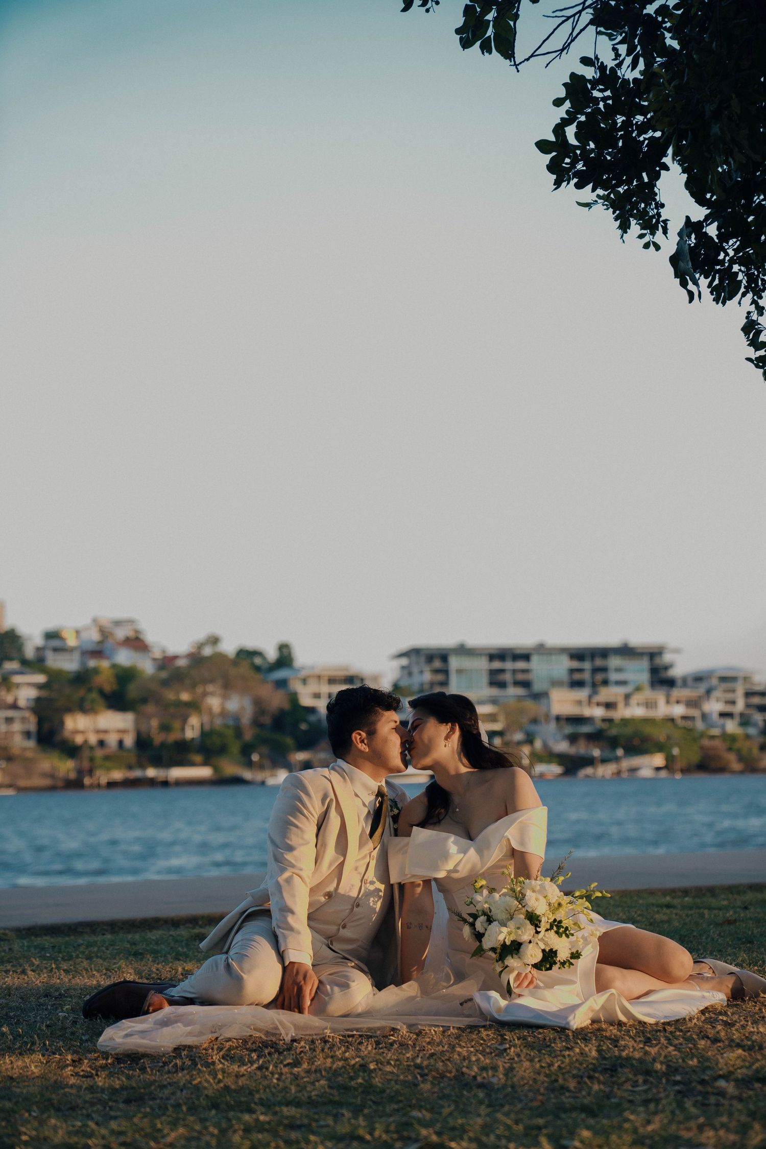 Romantic couple lounges together on beach at sunset with white floral bouquet and coastal buildings in background.