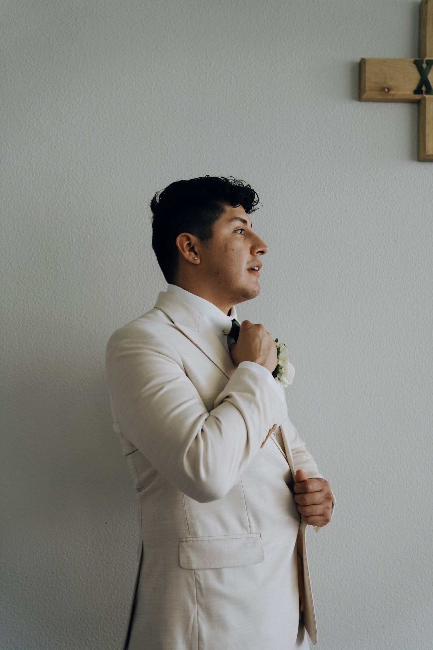 A person in a white suit jacket adjusts their tie while standing against a light-colored wall.