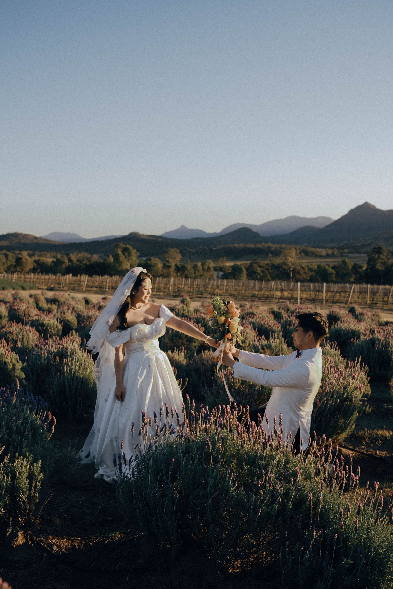 People in white dresses dance gracefully through a lavender field at sunset with mountains in the background.