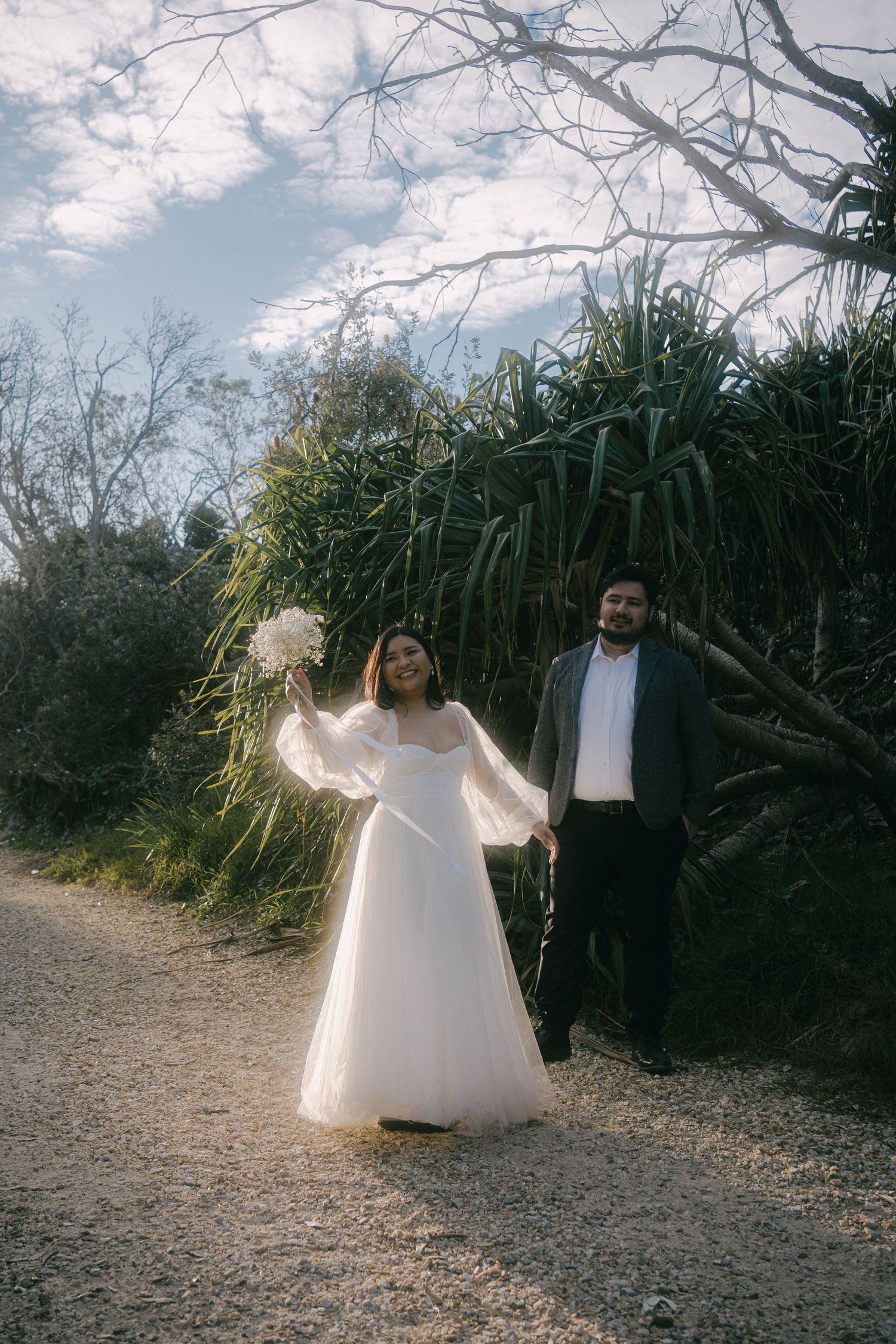 A couple poses together on a dirt path with tall grass and trees in the background during a wedding photoshoot.
