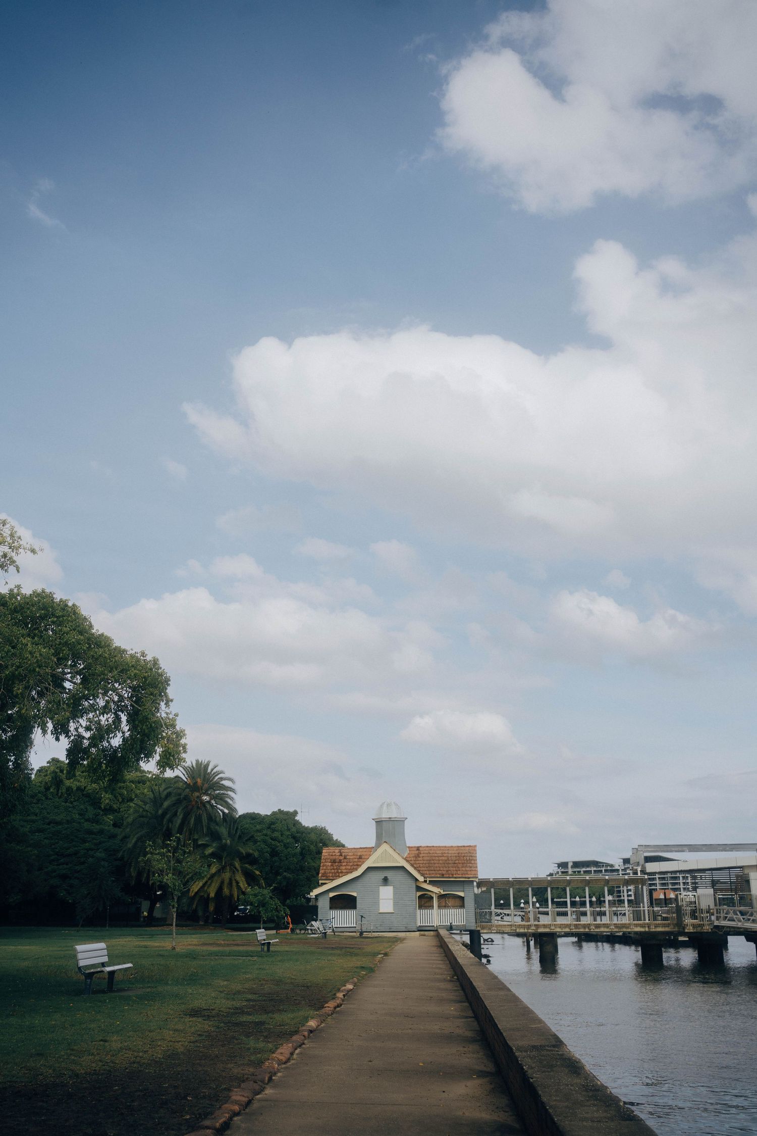 A waterfront walkway stretches along a coastal inlet with buildings and palm trees visible under partly cloudy skies.