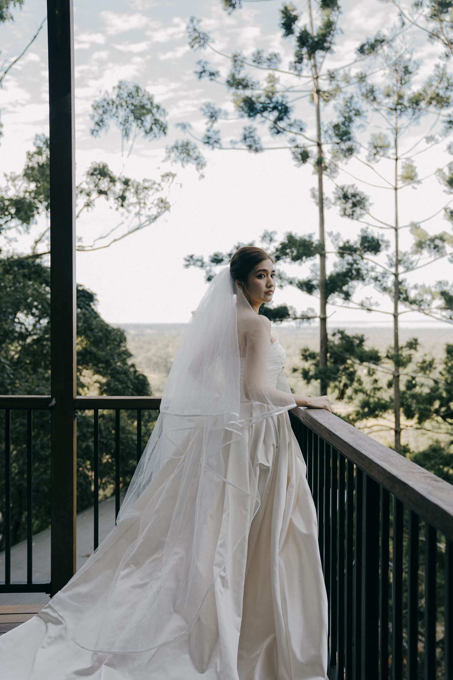 Ethereal wedding dress with long flowing veil billows gracefully on an outdoor balcony surrounded by pine trees.