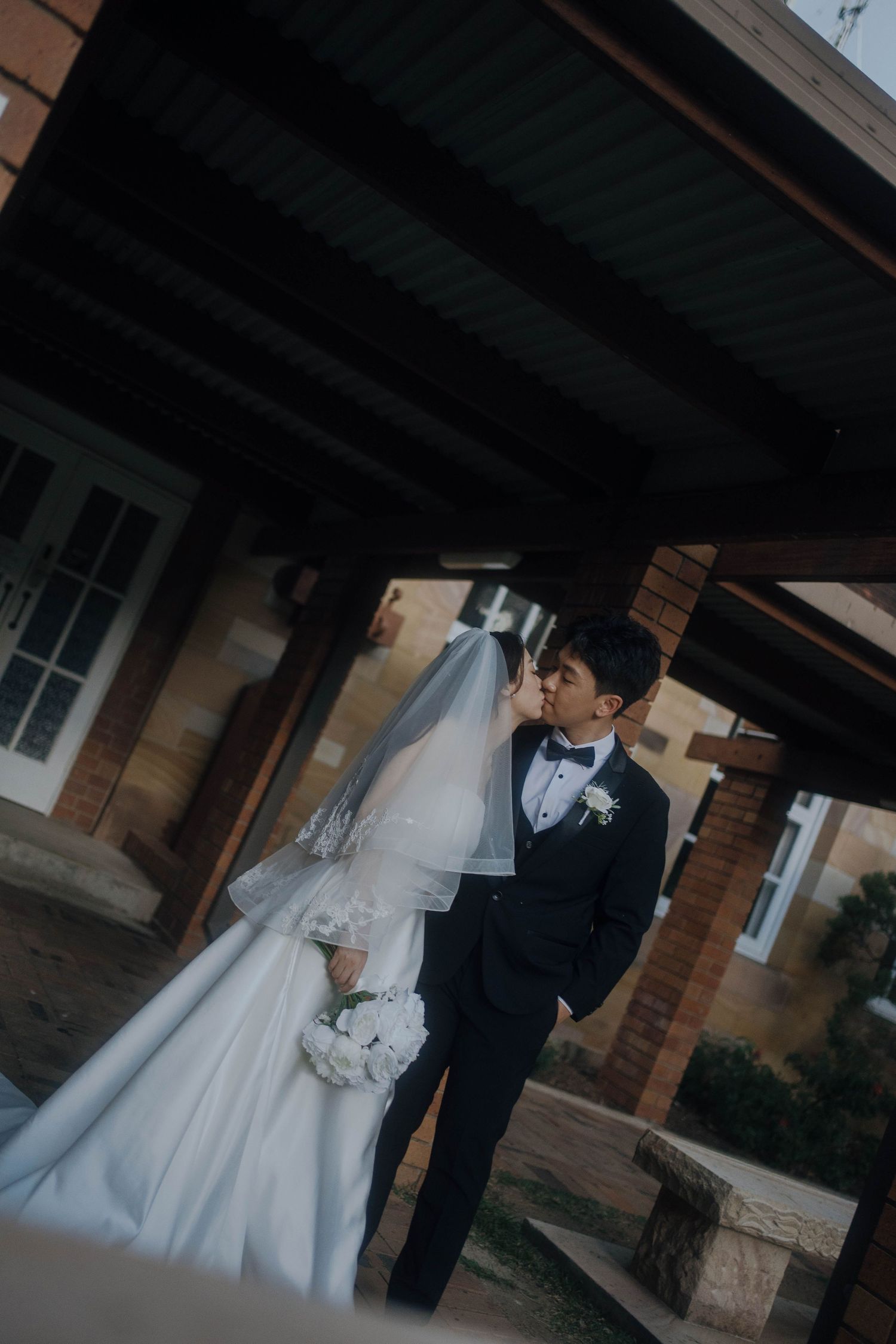 A romantic wedding couple shares an intimate moment under a covered porch of a brick building.