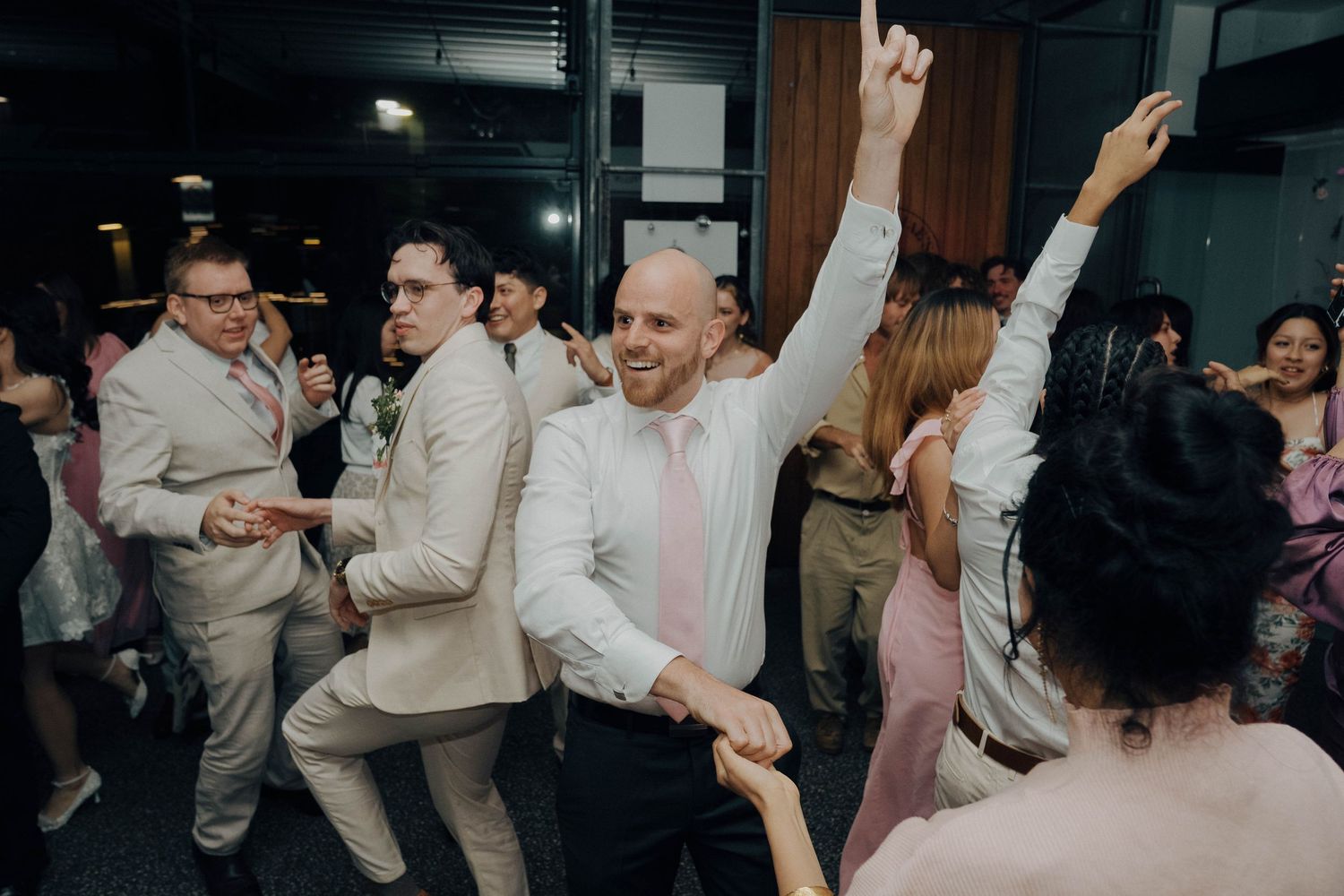 Wedding guests dance together on a crowded dance floor at an indoor reception venue.