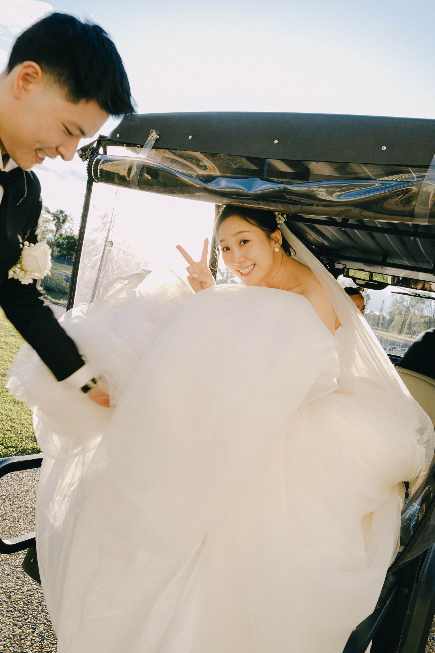 A bride in a flowing white wedding dress is being helped into a black car by her companion.