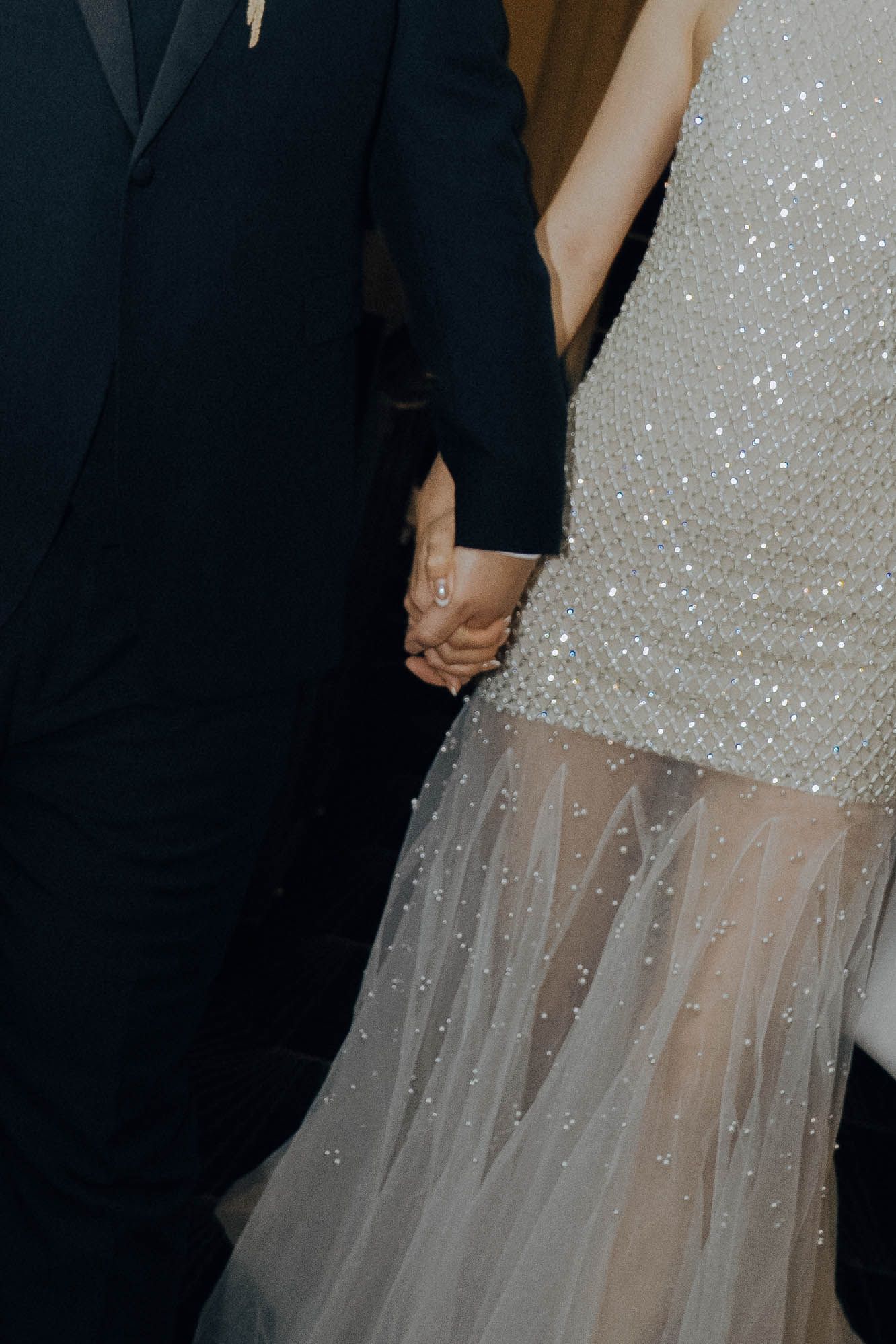 Close-up detail of a sparkly white wedding dress with sheer tulle overlay next to a dark suit at formal event.