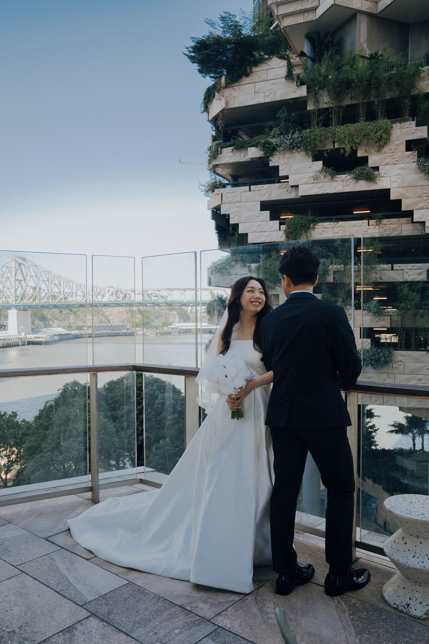A couple in wedding attire stands on a balcony overlooking a river and bridge in an architectural setting.