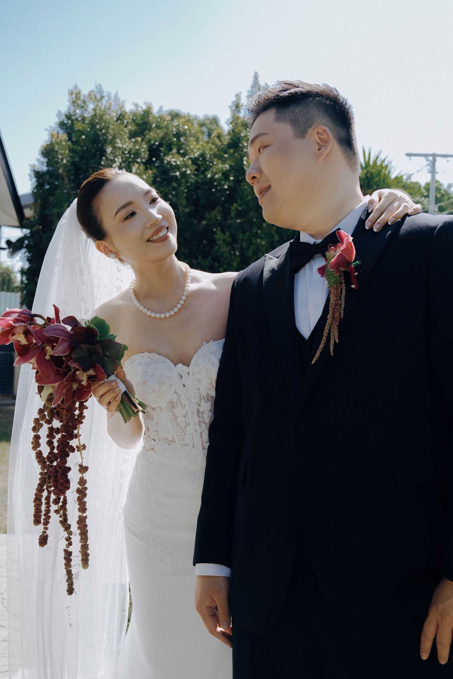 A couple in wedding attire share a joyful moment outdoors, with the bride holding a cascading burgundy floral bouquet.