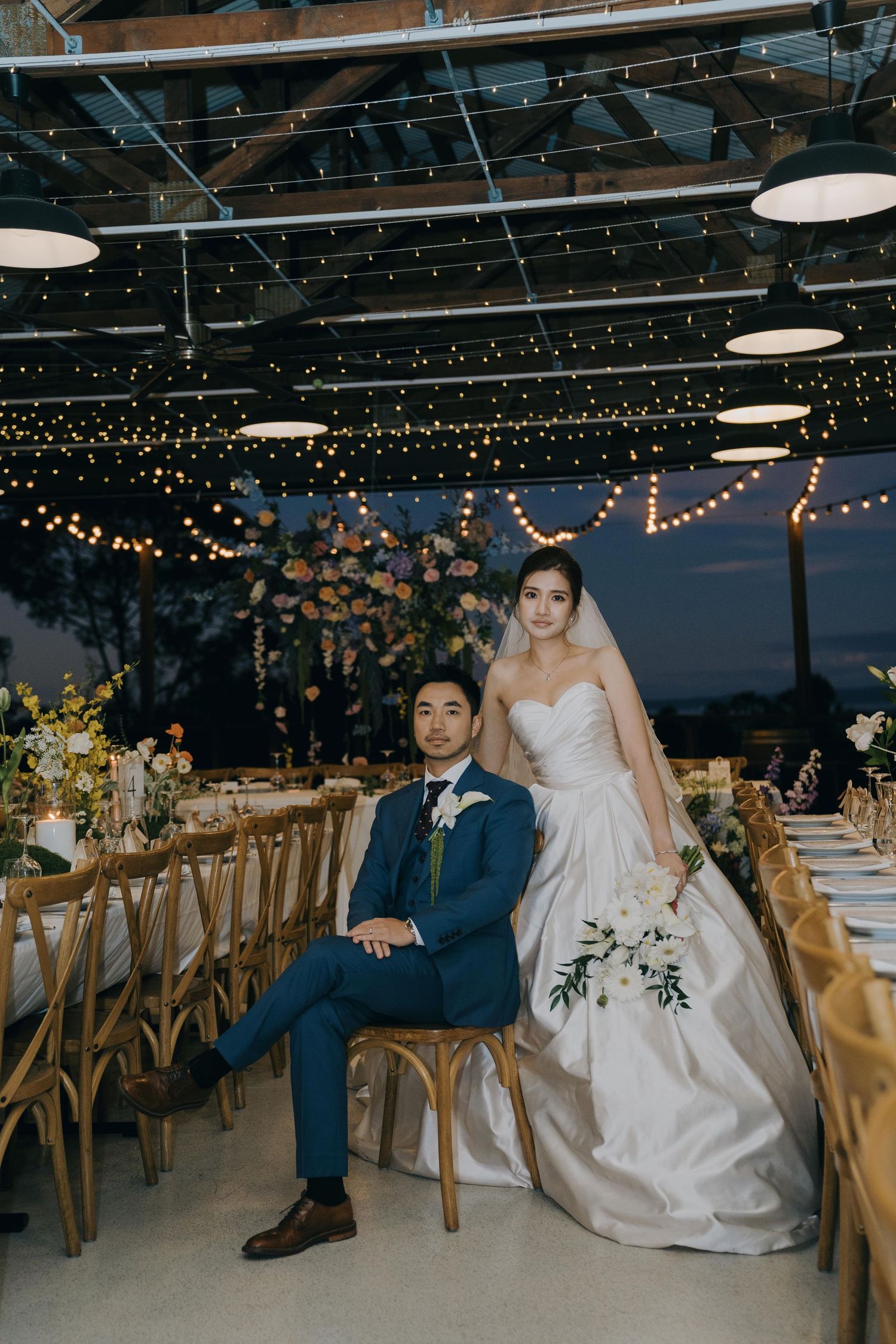 Indoor wedding reception venue decorated with string lights and wooden chairs with couple seated at dining table.
