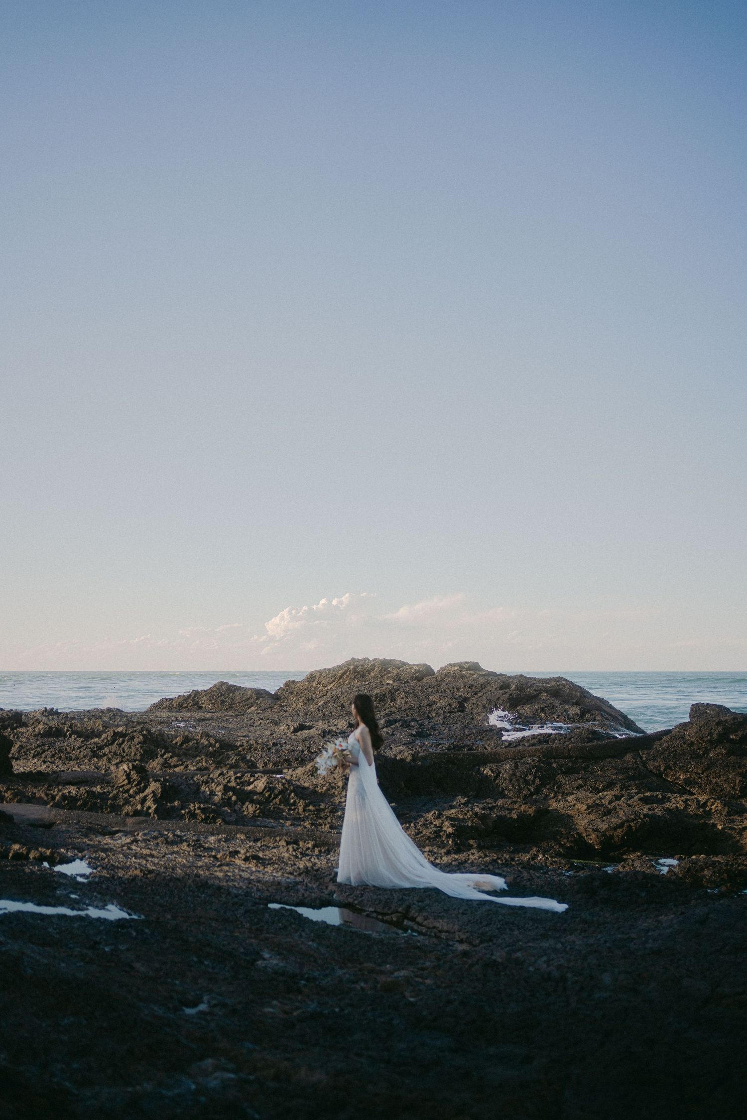 A figure in a flowing white wedding dress stands gracefully against rocky coastal cliffs at sunset by the ocean.