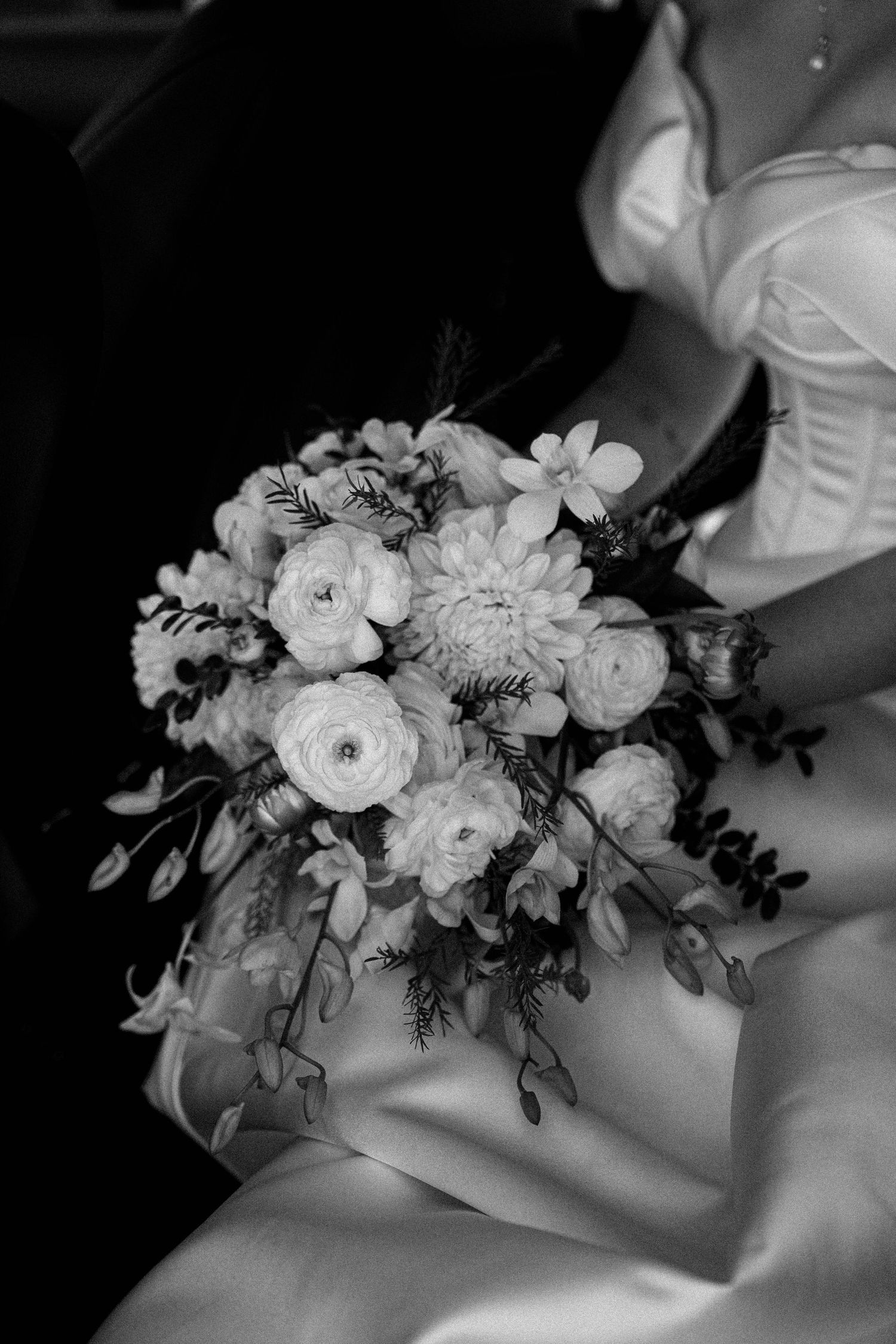 Black and white wedding photo showing elegant floral bouquet with ranunculus and greenery against white fabric.