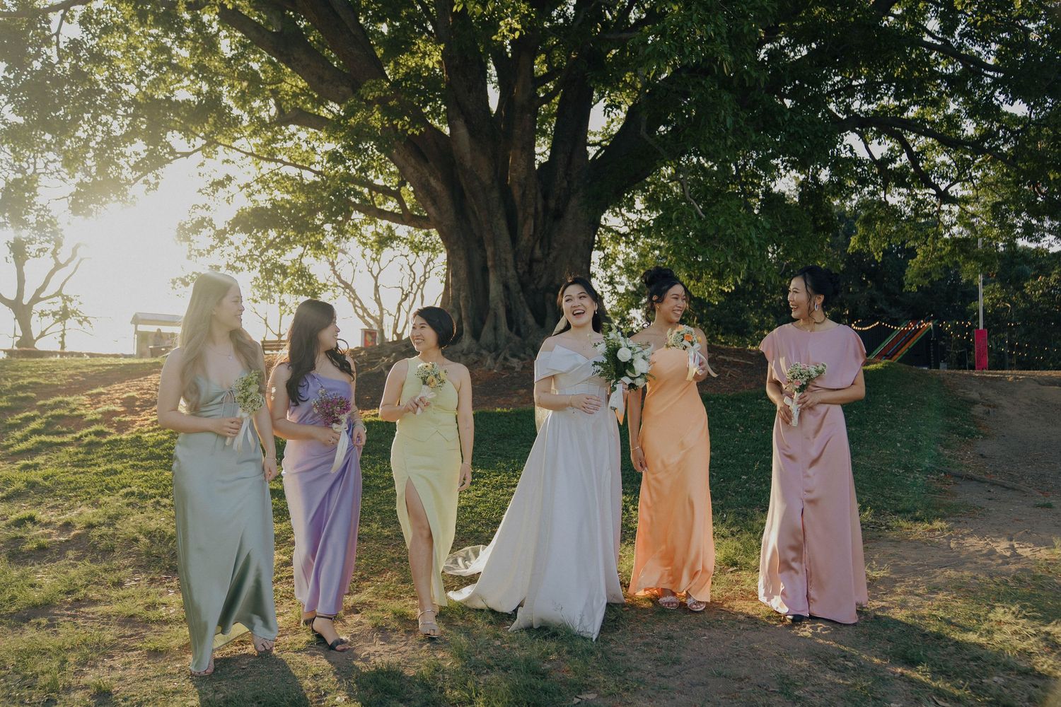A wedding party in pastel purple dresses stands together under trees during golden hour sunset.