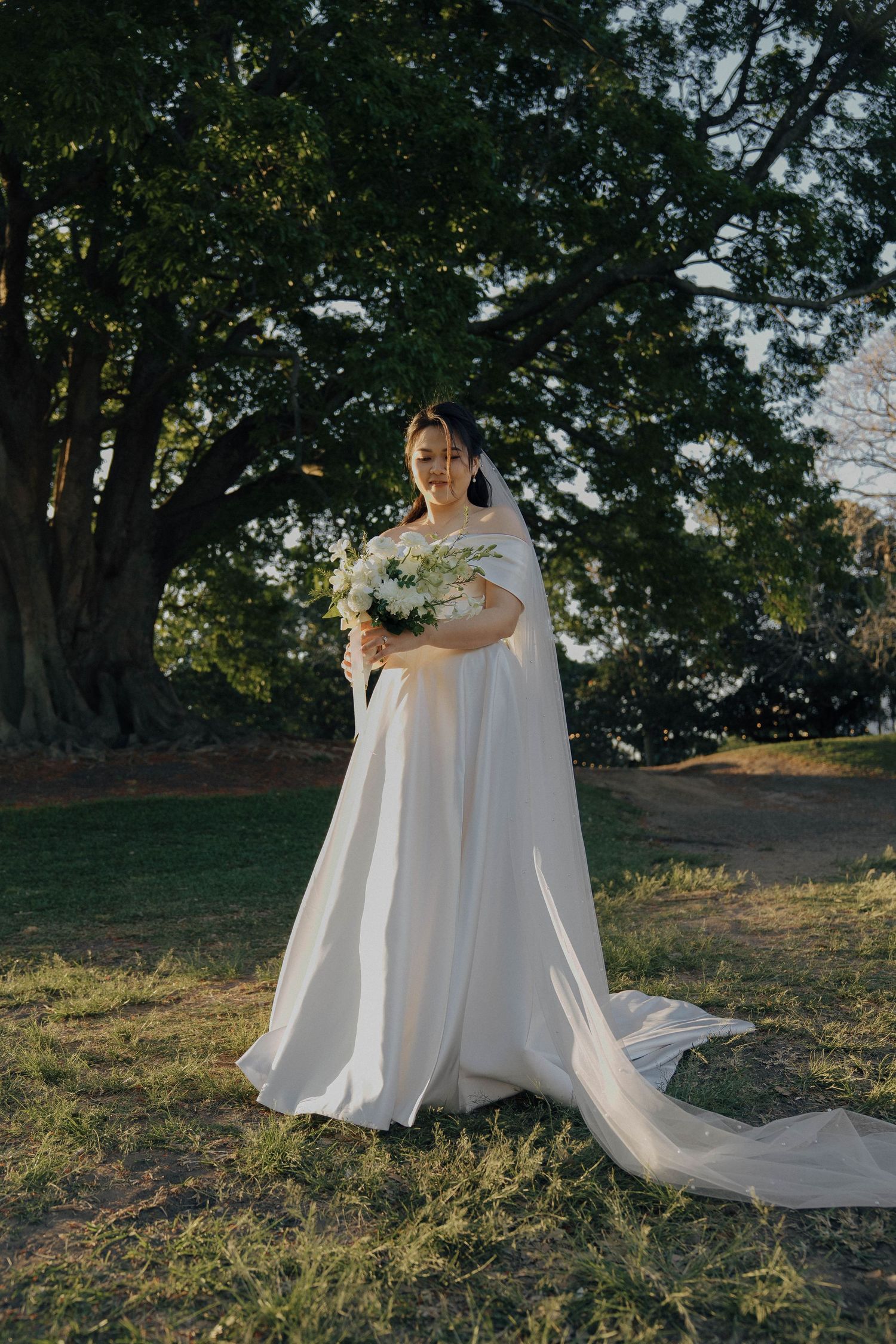 A flowing white wedding dress with a long train cascades across green grass in a sunlit outdoor setting.