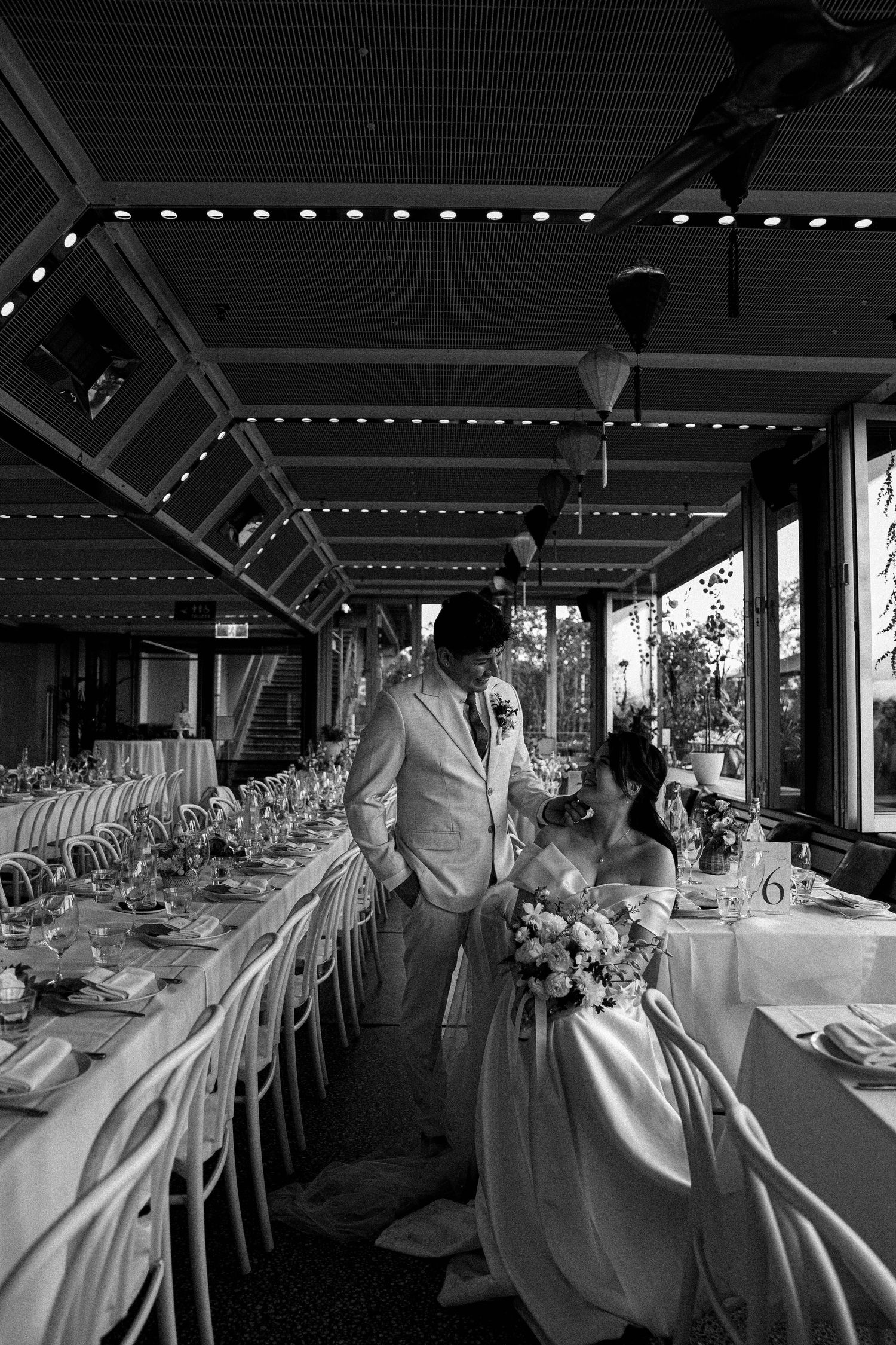 Black and white panoramic view of an elegant wedding reception venue with long tables and decorative lighting.