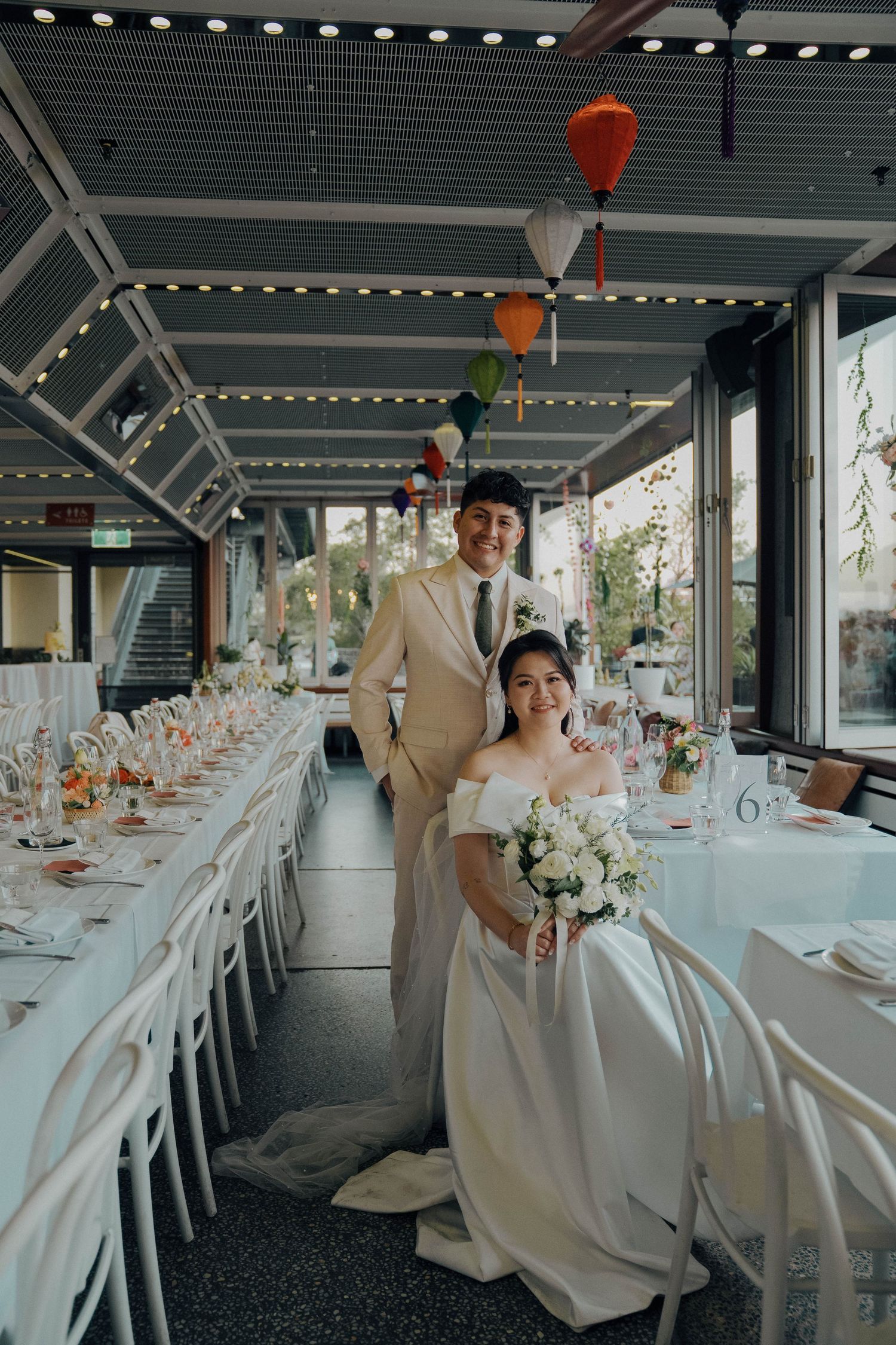 Elegant wedding reception venue with white chairs, long tables, and hanging lanterns in an industrial-modern space.