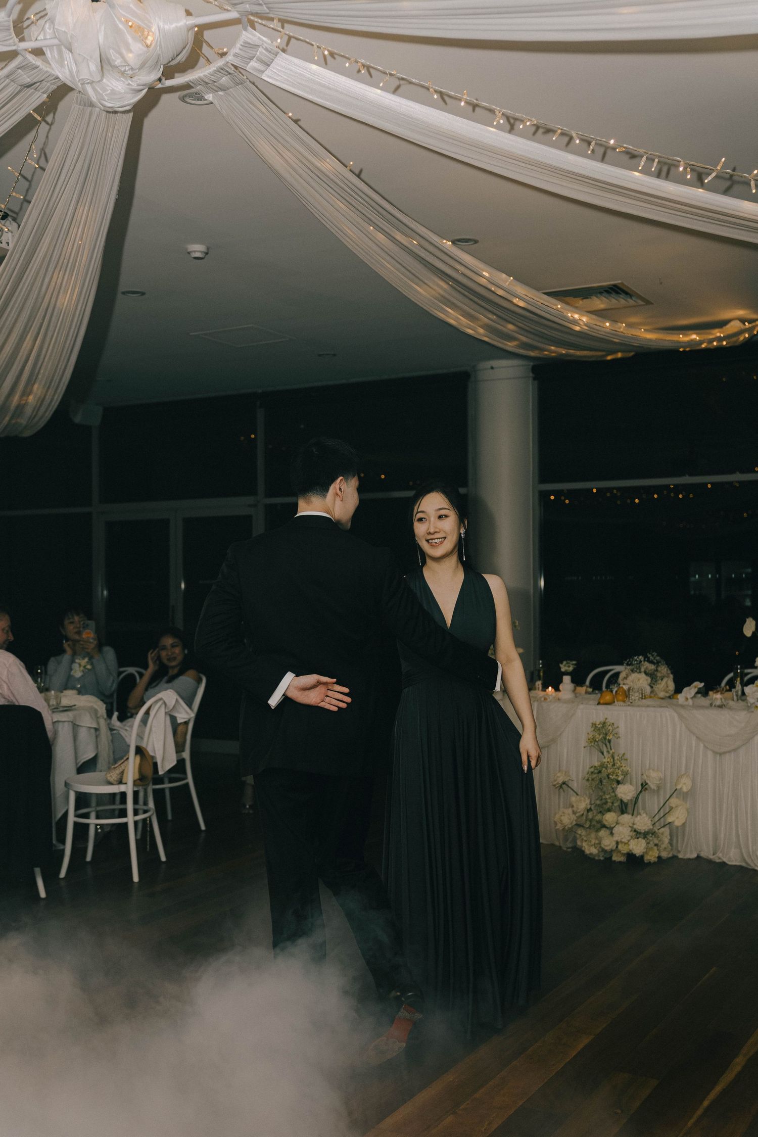 Couples dancing on a dark dance floor with white fabric draped decor and atmospheric fog effects at an elegant event.