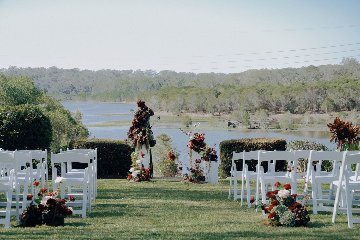 Panoramic view of white wedding chairs set up on a grassy hillside overlooking a lake with floral arrangements.