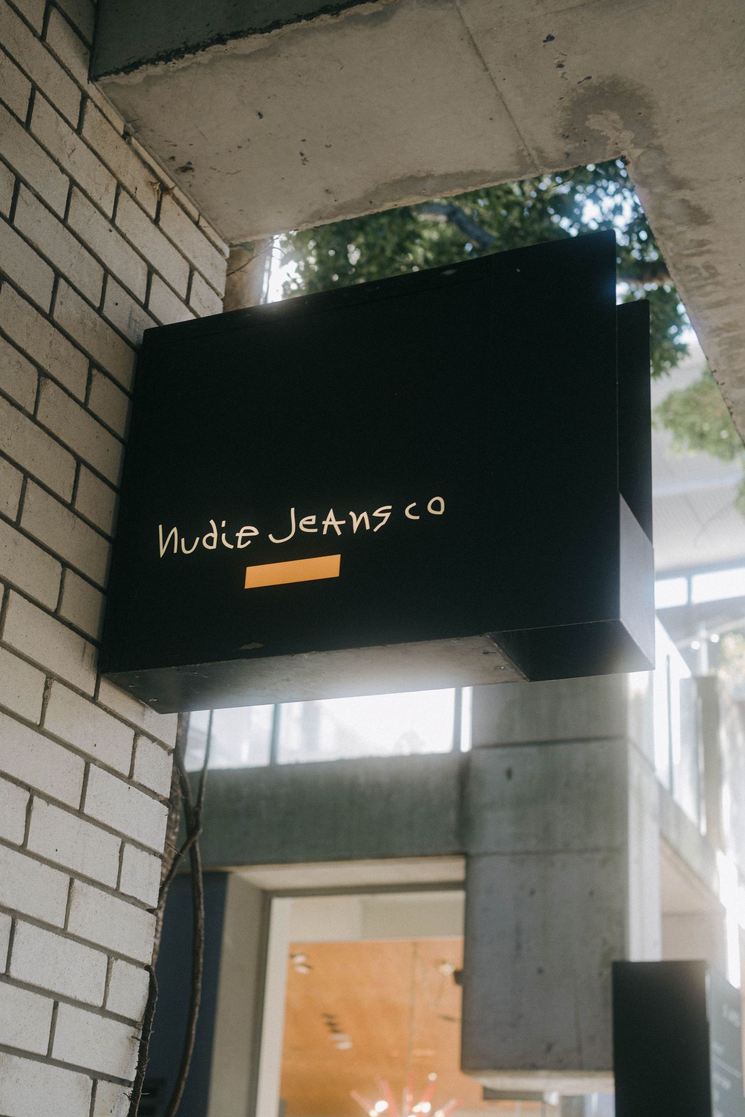 Black storefront sign for Nudie Jeans Co mounted on white brick wall exterior.