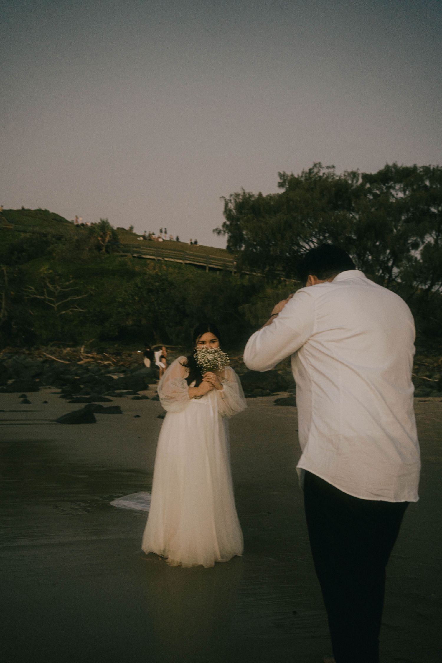 A photographer captures a couple in wedding attire on a moody beach with dark trees in the background.