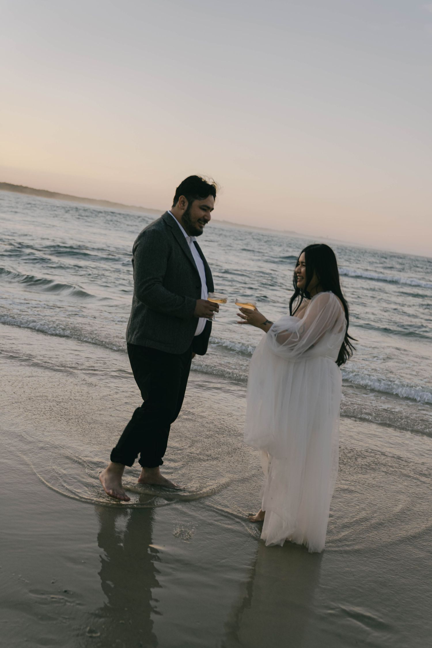 A couple stands together on a beach at sunset, with waves lapping at their feet as they share an intimate moment.