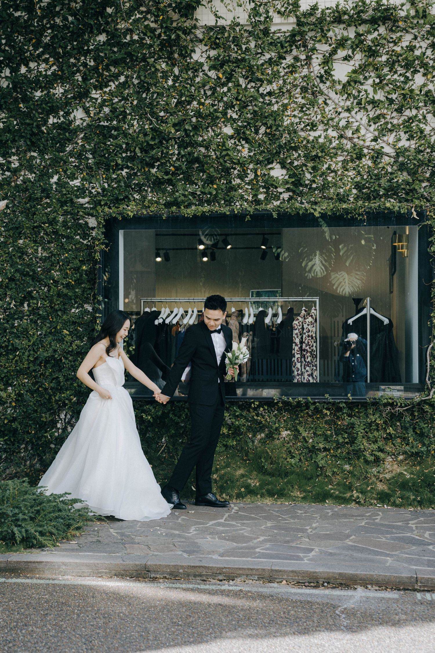 A couple in formal wedding attire dances together on a stone patio surrounded by lush greenery and trees.