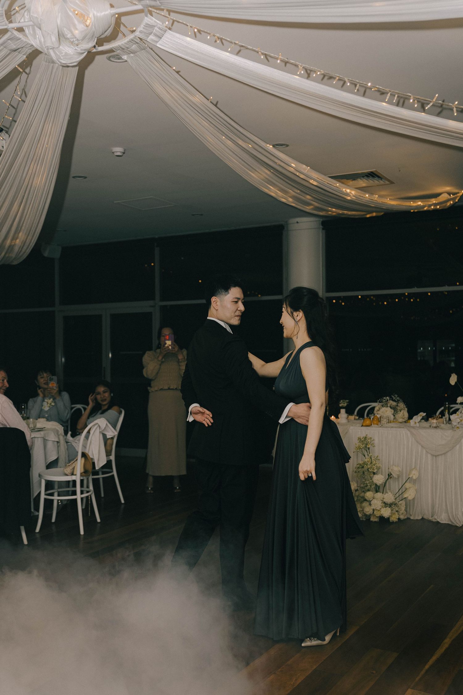 Couples dance on a dimly lit ballroom floor with white draped decor and fog effects creating an elegant atmosphere.