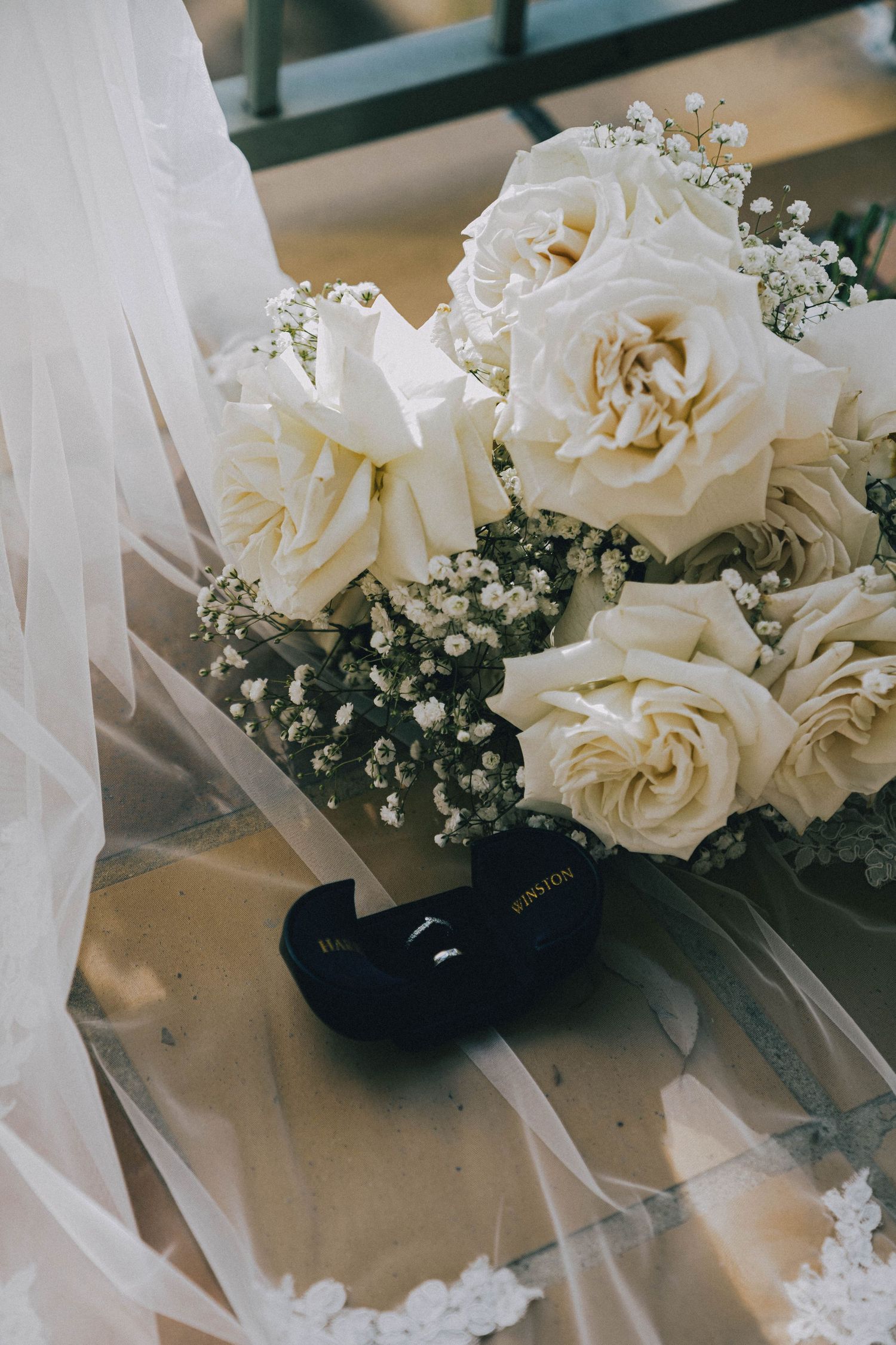 A delicate bridal bouquet of white roses and baby's breath rests on sheer tulle veil next to black wedding rings.
