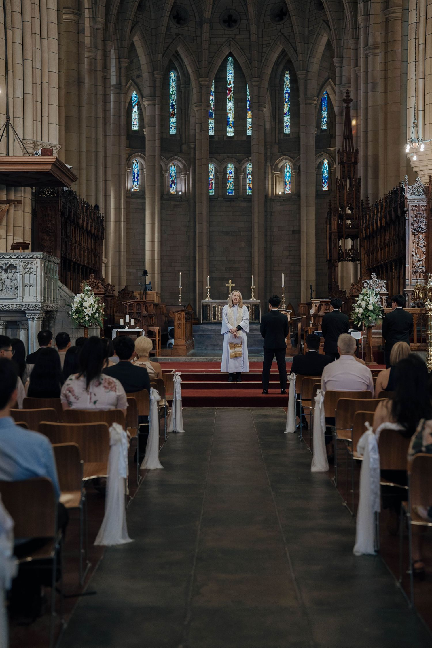 Interior view of a Gothic cathedral during a wedding ceremony with white floral arrangements along the aisle.