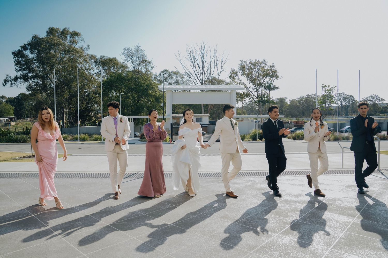Wedding party members walking in a line cast long shadows on a sunny day at an outdoor venue.