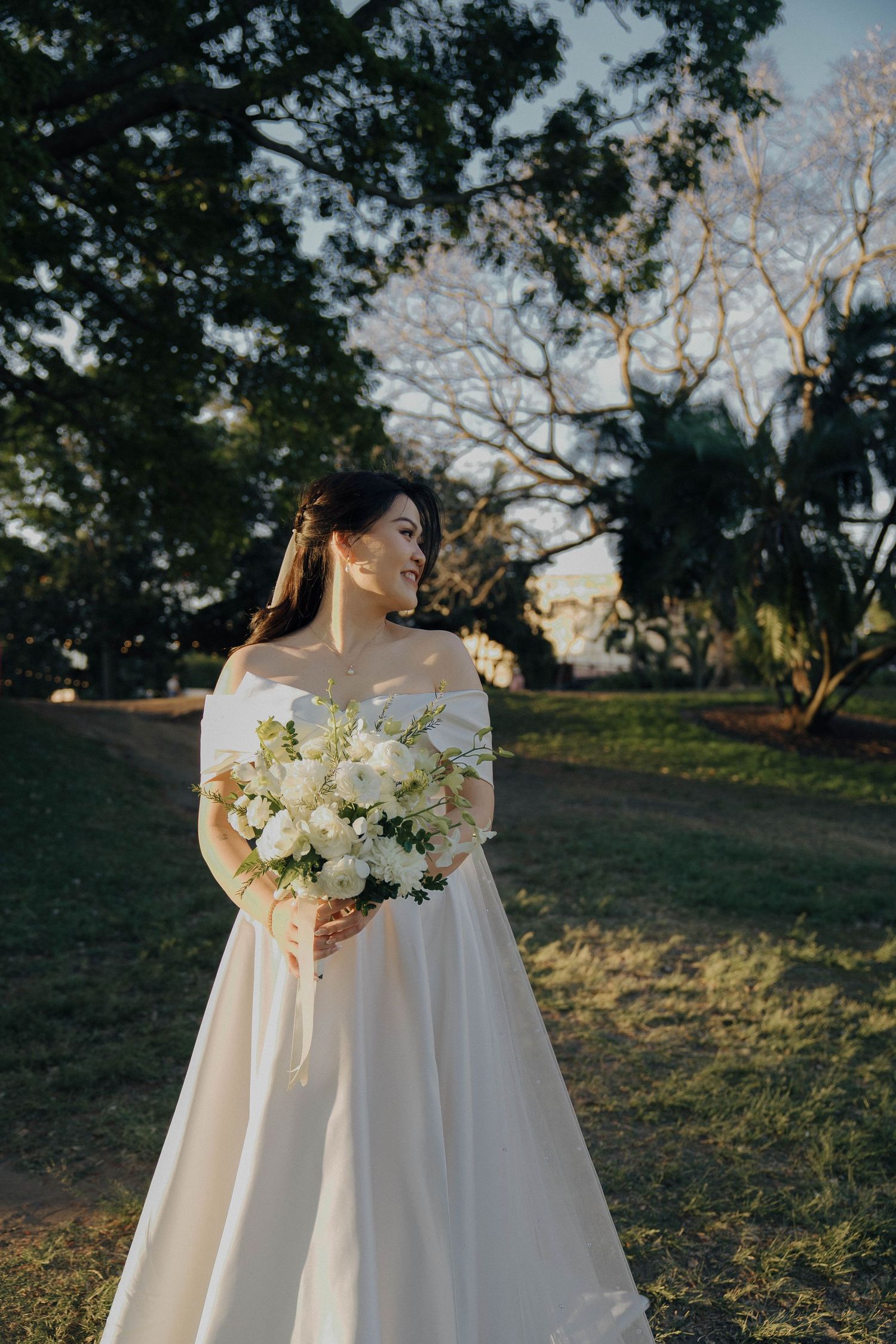 A bride in a flowing white dress poses with a white floral bouquet against a natural outdoor backdrop at sunset.