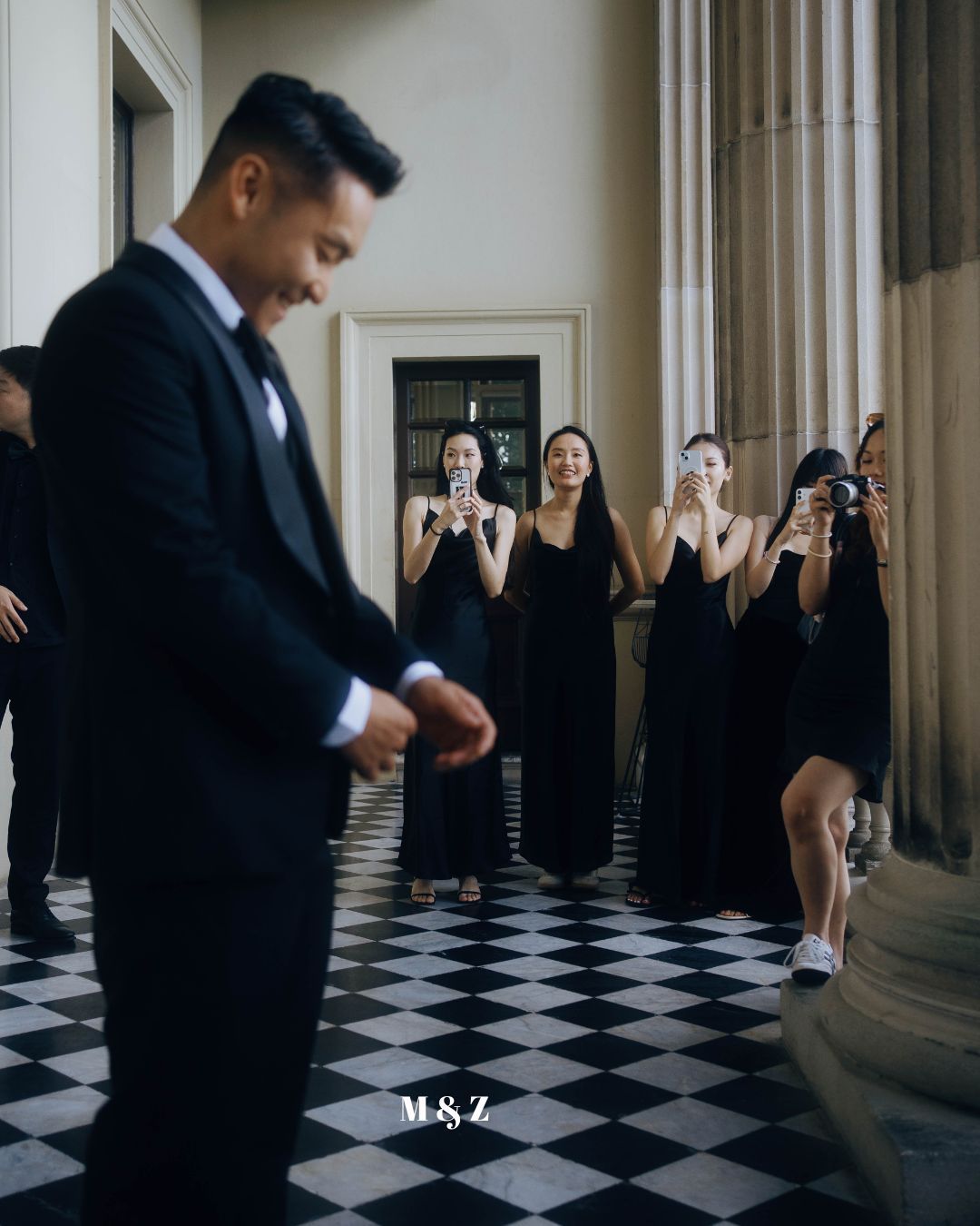 A person in a black suit checks their watch while others in black dresses hold cameras in a checkered floor hallway.