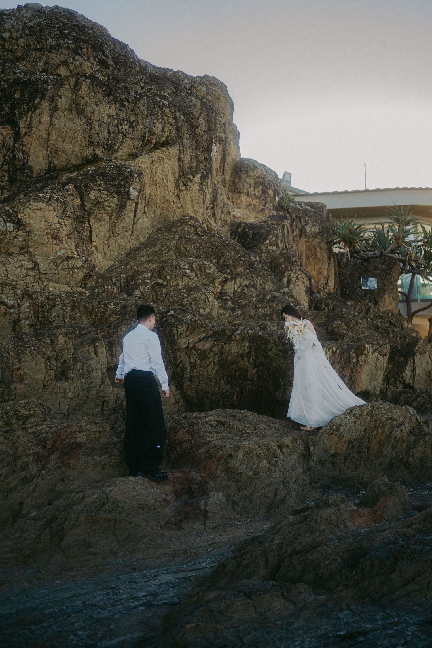 Two figures stand among rocky coastal cliffs at dusk, one wearing a white dress as they explore the rugged terrain.
