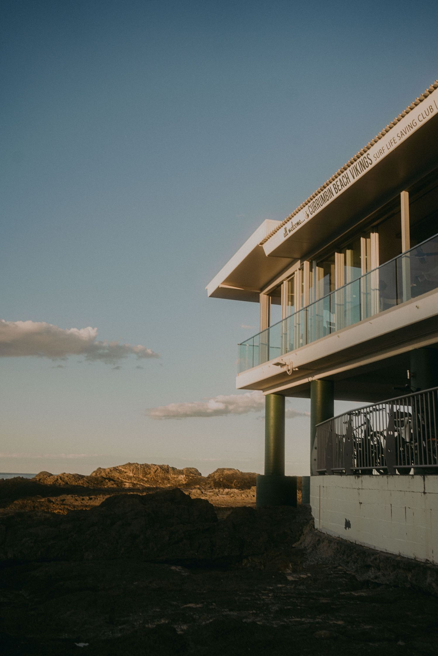 A modern minimalist desert house with large glass railings and clean white walls sits against a mountainous landscape at sunset.