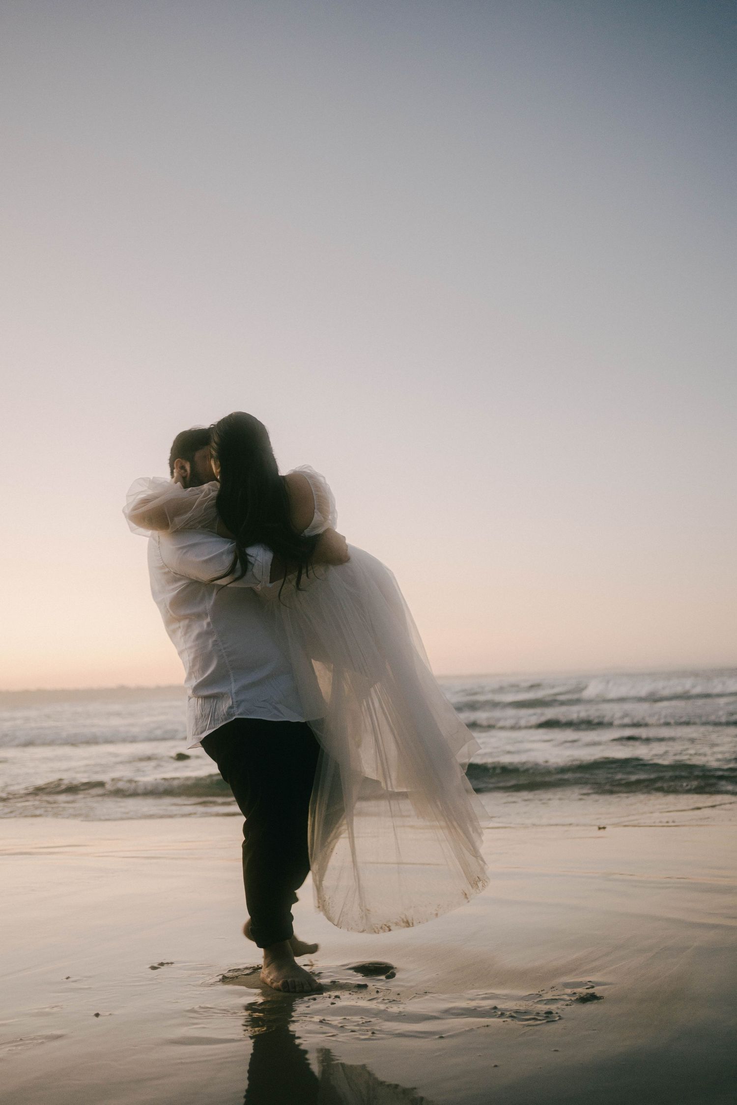 A couple embraces on a misty beach at sunset as waves crash in the background.