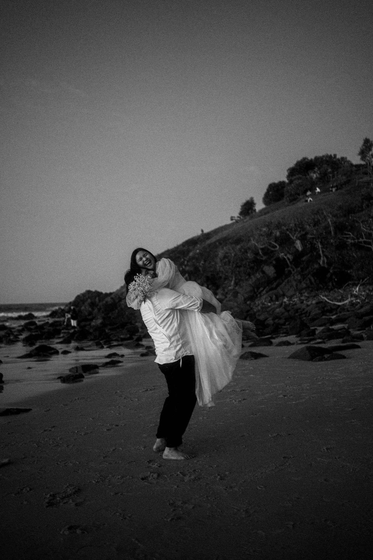 A romantic black and white photo of a couple embracing on a rocky beach with hills in the background.