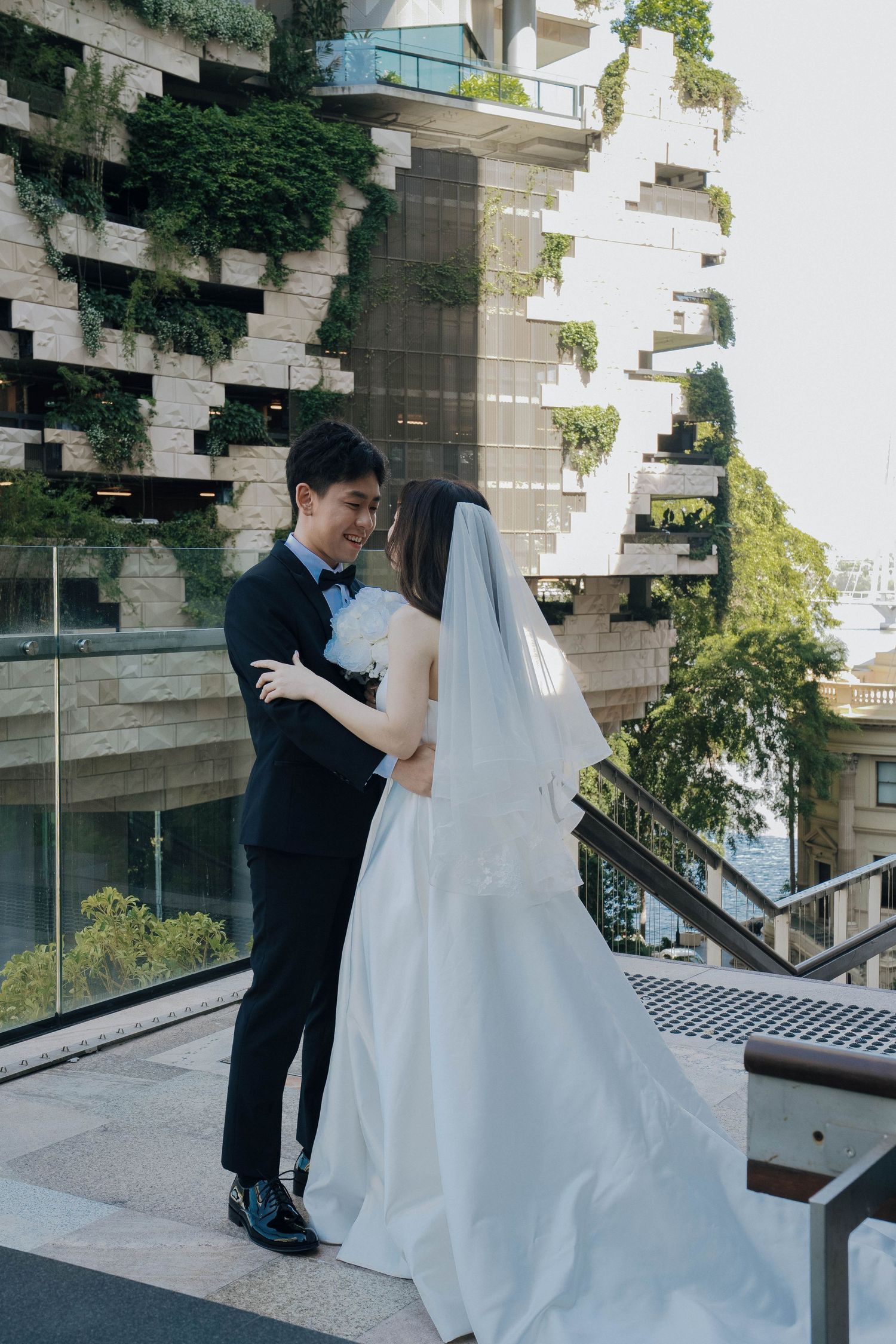 A couple in wedding attire shares a romantic moment on outdoor steps of a modern building.