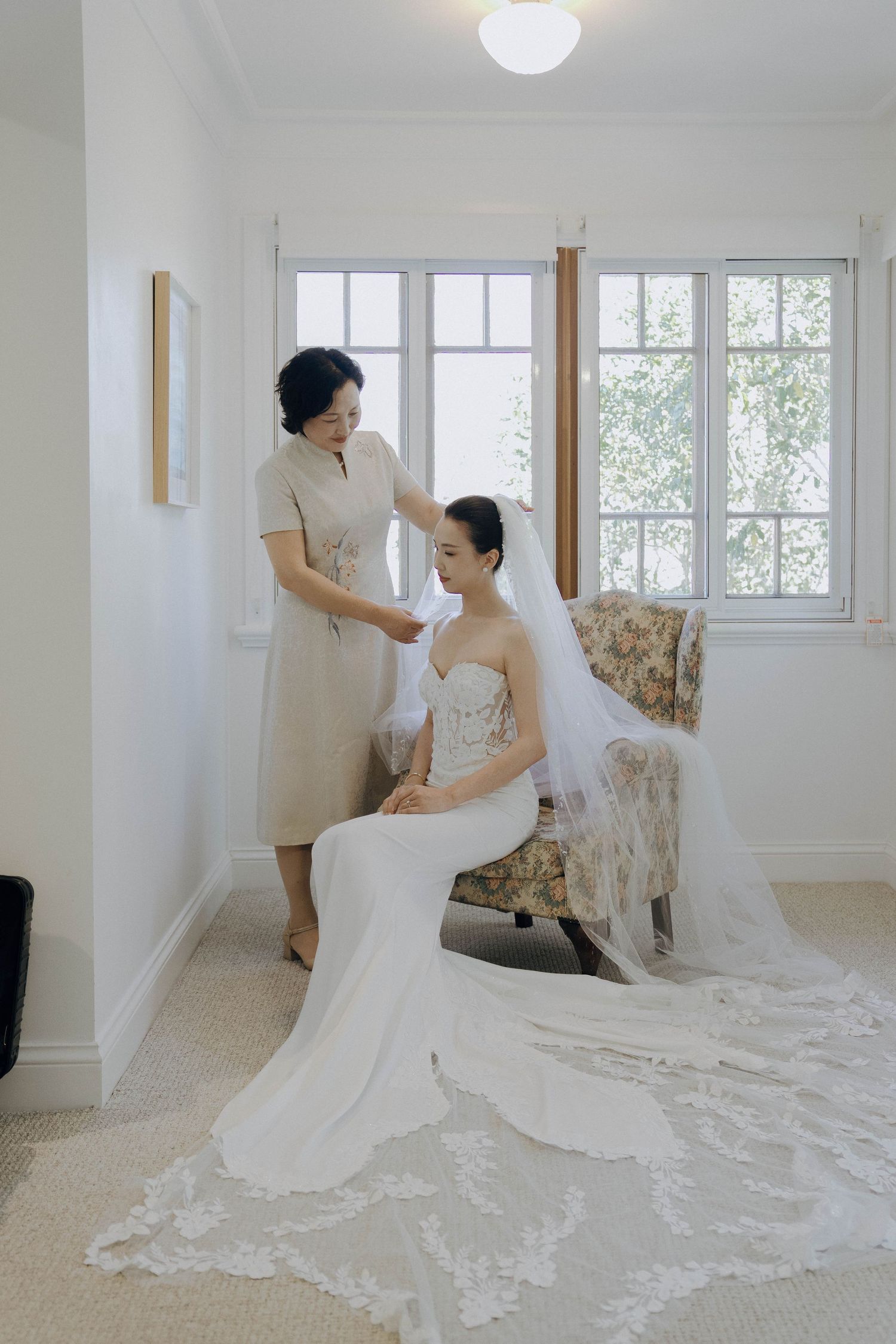 A bride in a fitted white wedding gown with long lace veil gets final adjustments in a bright sunlit room.