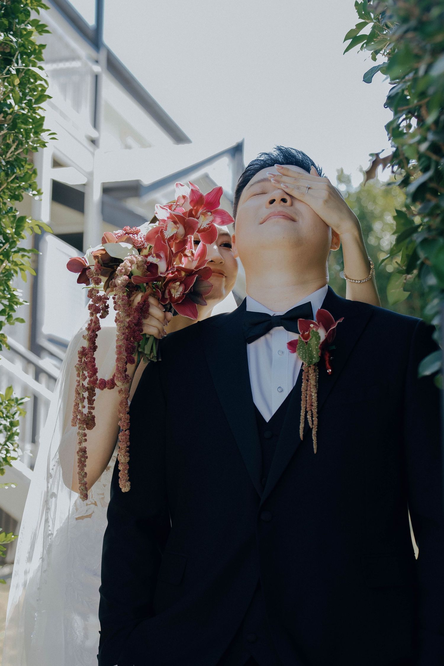 A person in a formal black tuxedo and bowtie holding pink flowers stands against a white building exterior.