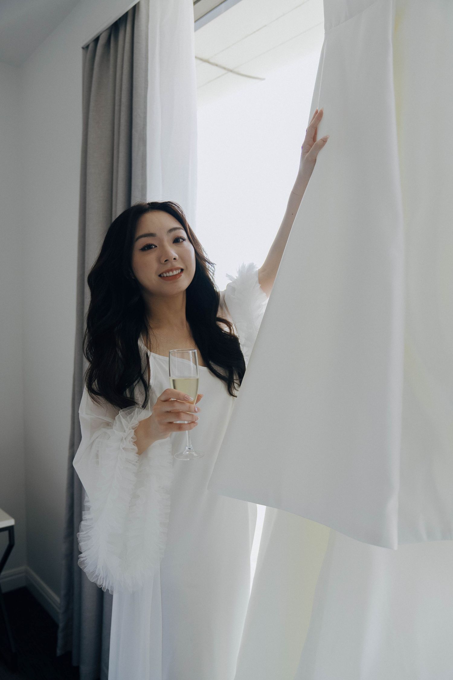A person in white clothing dances joyfully near sheer curtains in a bright sunlit room.