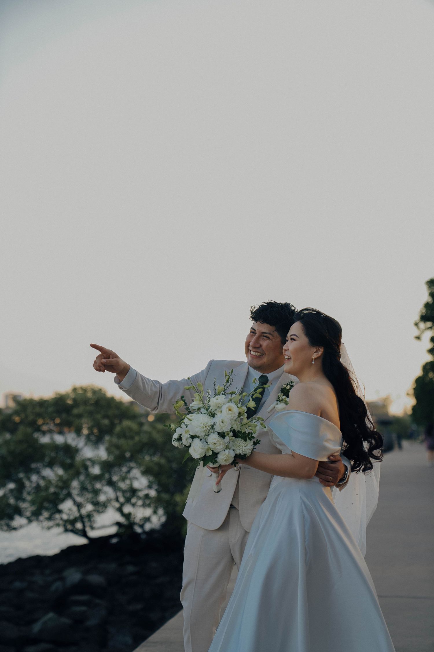 A couple in wedding attire share a joyful moment together at sunset while pointing into the distance.