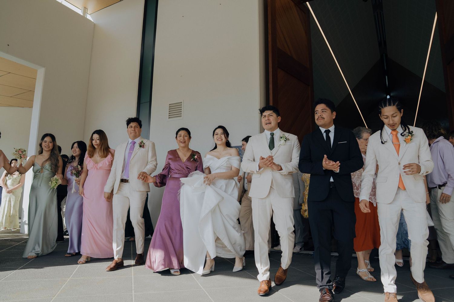 Wedding party members in formal attire including white suits and pastel bridesmaid dresses line up against a wall.
